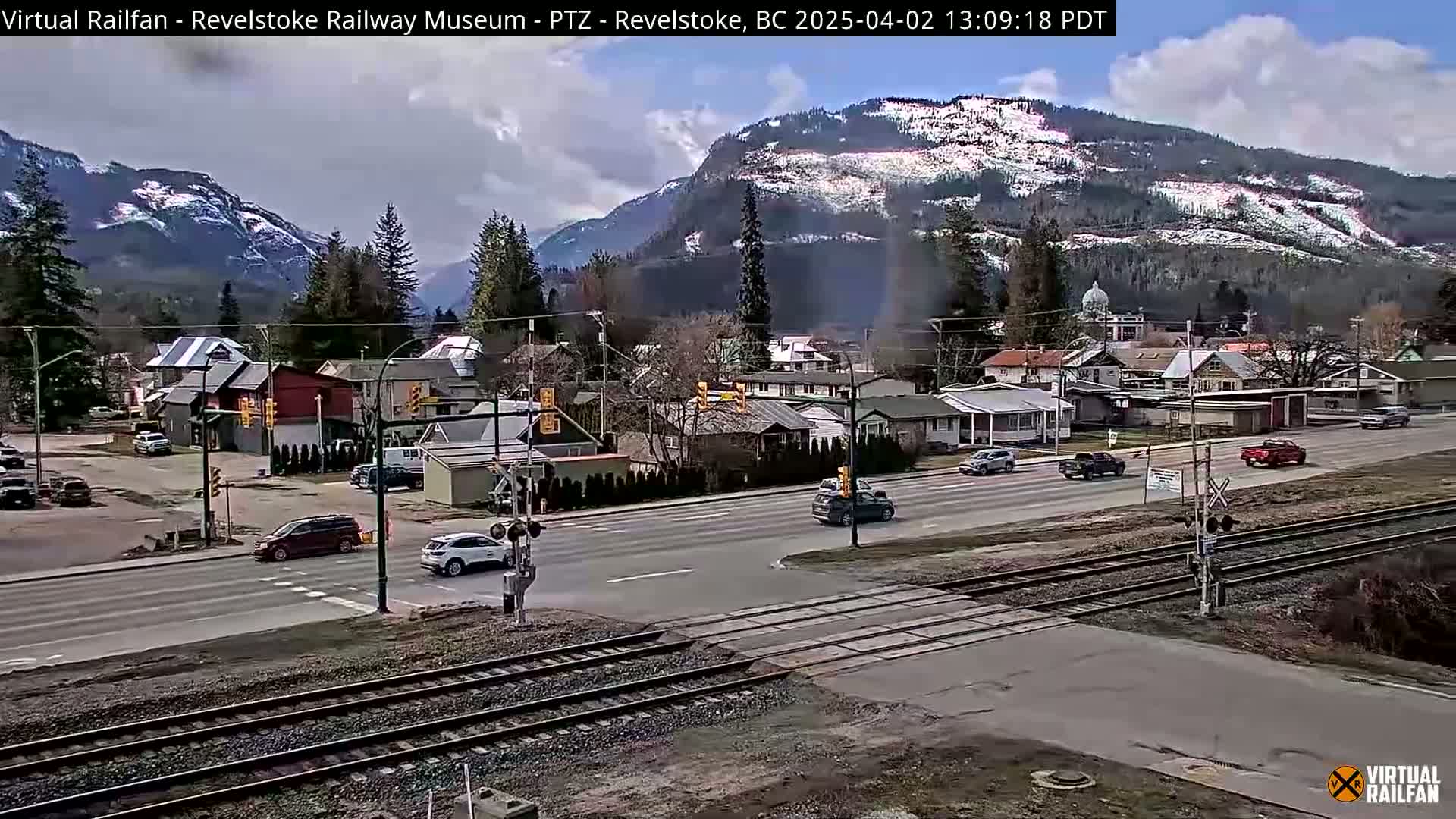 A partly cloudy day reveals a small town nestled in a valley, with snow-capped mountains in the background, and several cars driving along a road that crosses railroad tracks.