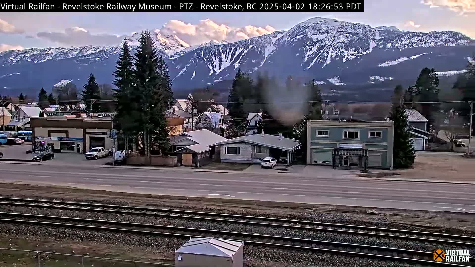 A partly cloudy evening view of a town with snow-capped mountains in the background, railroad tracks in the foreground, and a few vehicles parked along the road.