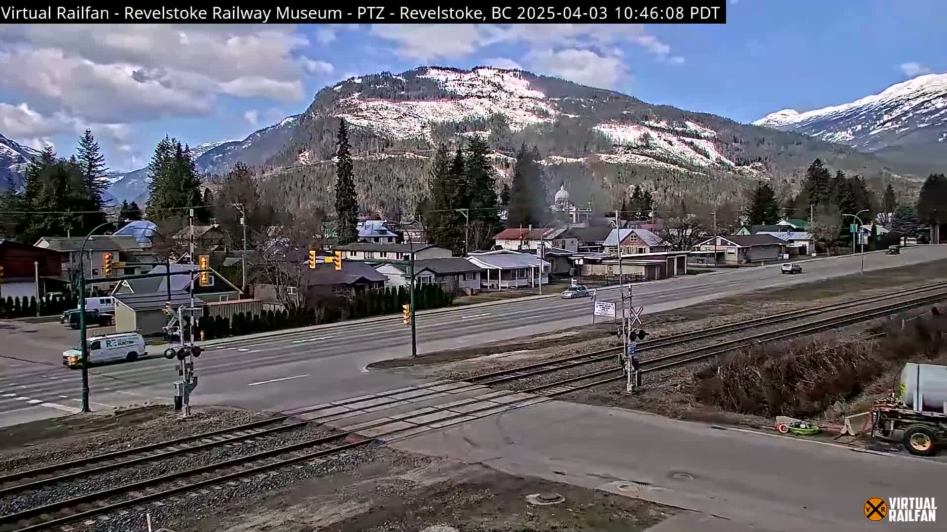 A partly sunny day reveals a town nestled in a valley between snow-capped mountains, with a road and railway tracks intersecting in the foreground.