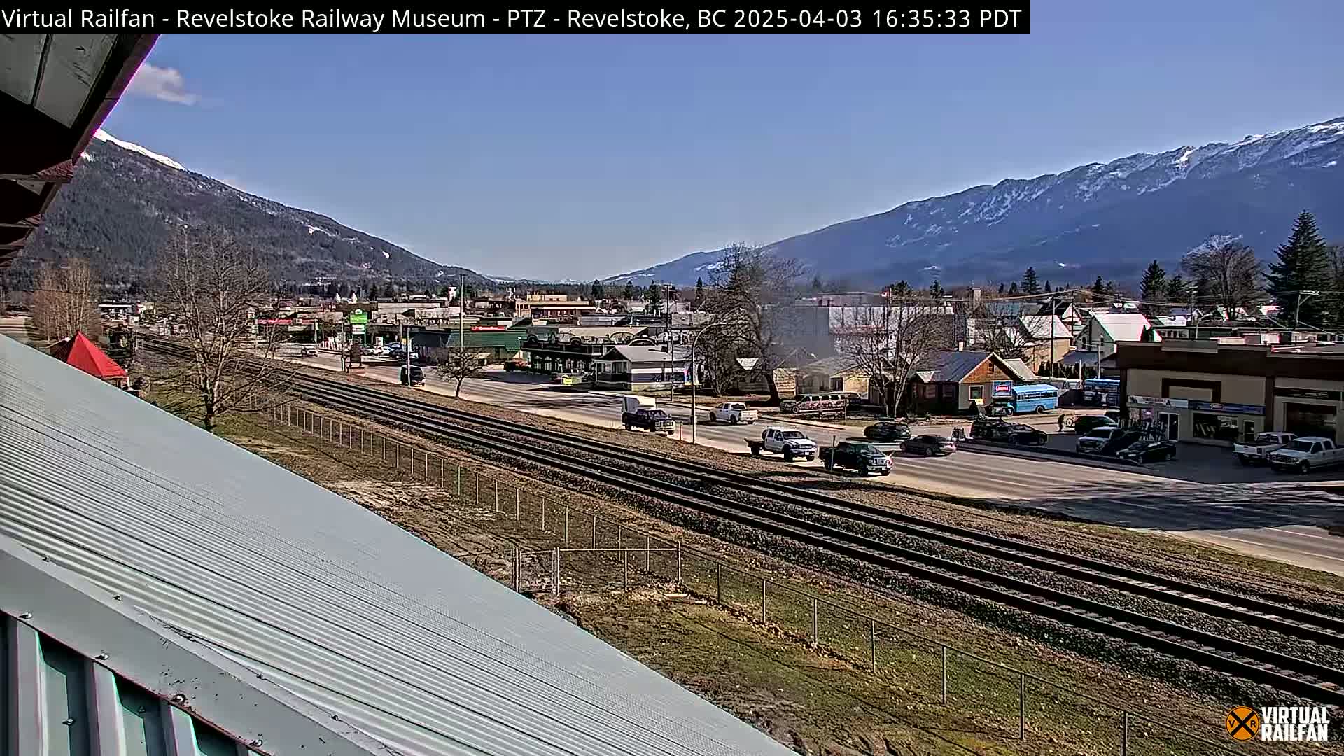 A sunny view of a town nestled in a valley between mountains, with train tracks running alongside a road, and a few vehicles visible.