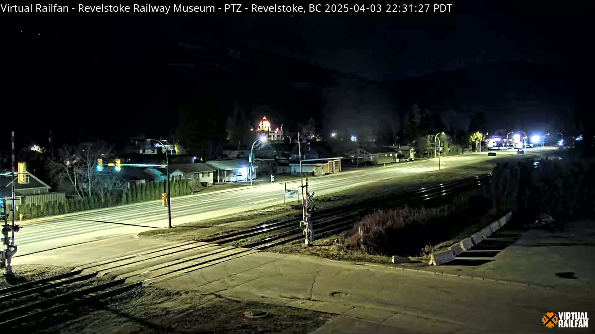 A nighttime view of a town's street and railway crossing, illuminated by streetlights, with a mountain range silhouetted against a dark sky.