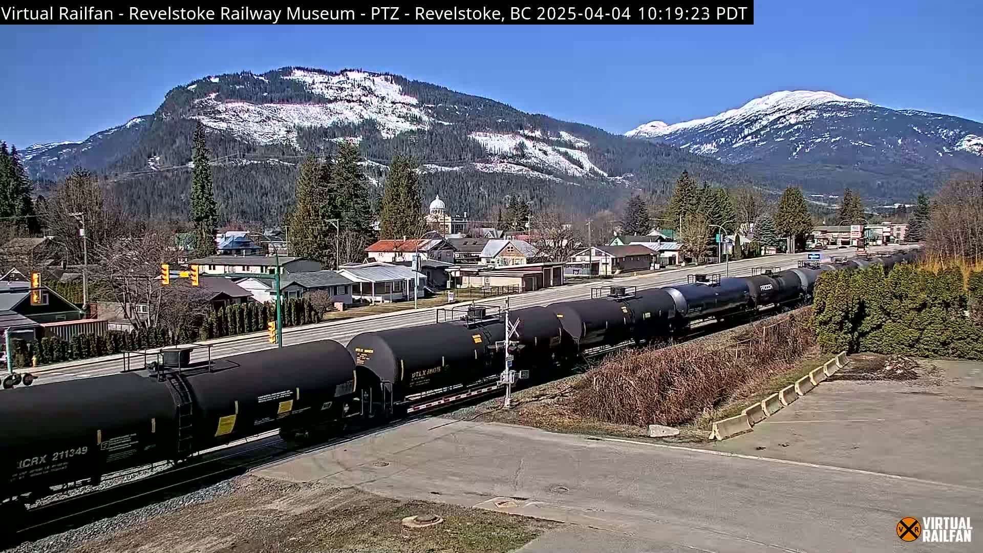 A long train of black tanker cars is passing through a small town nestled in a valley surrounded by snow-capped mountains under a sunny sky.