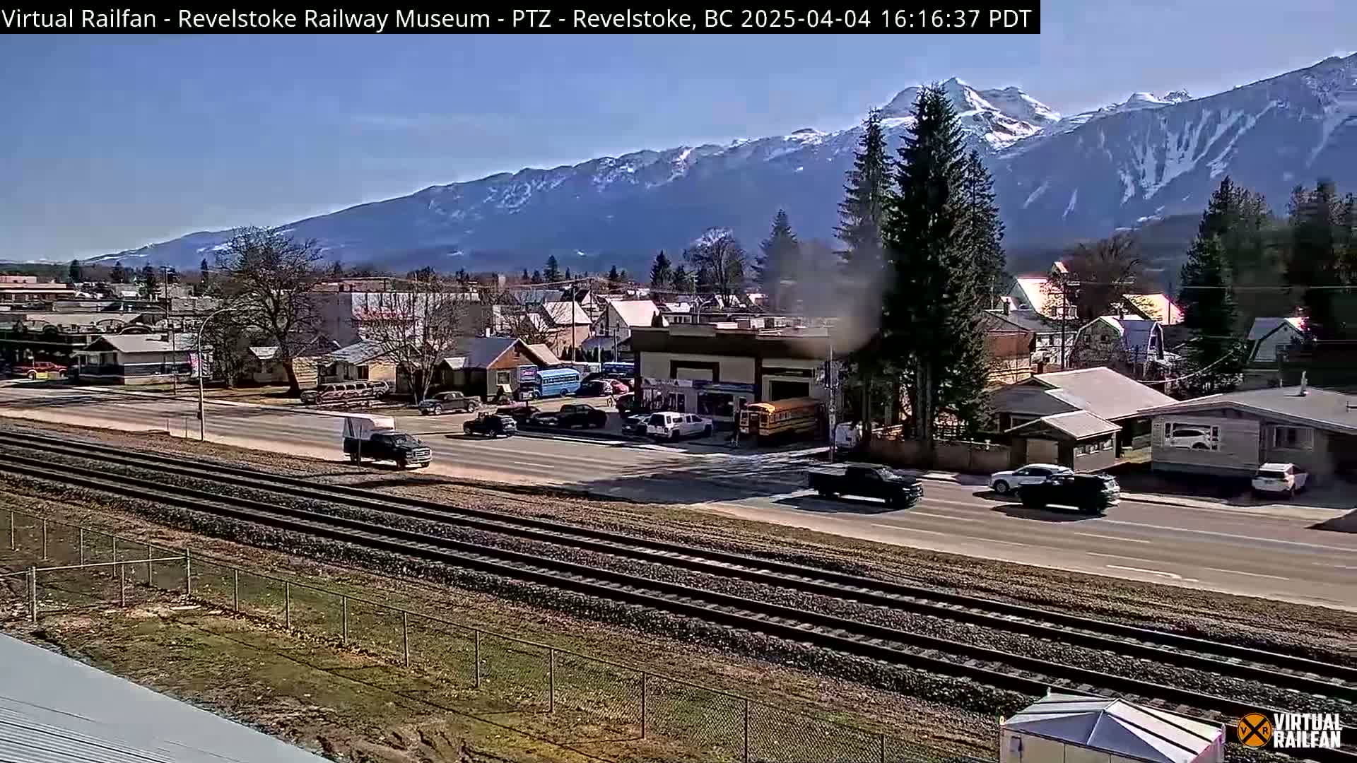 A sunny day reveals a small town nestled in a valley, with snow-capped mountains in the background and railway tracks in the foreground.