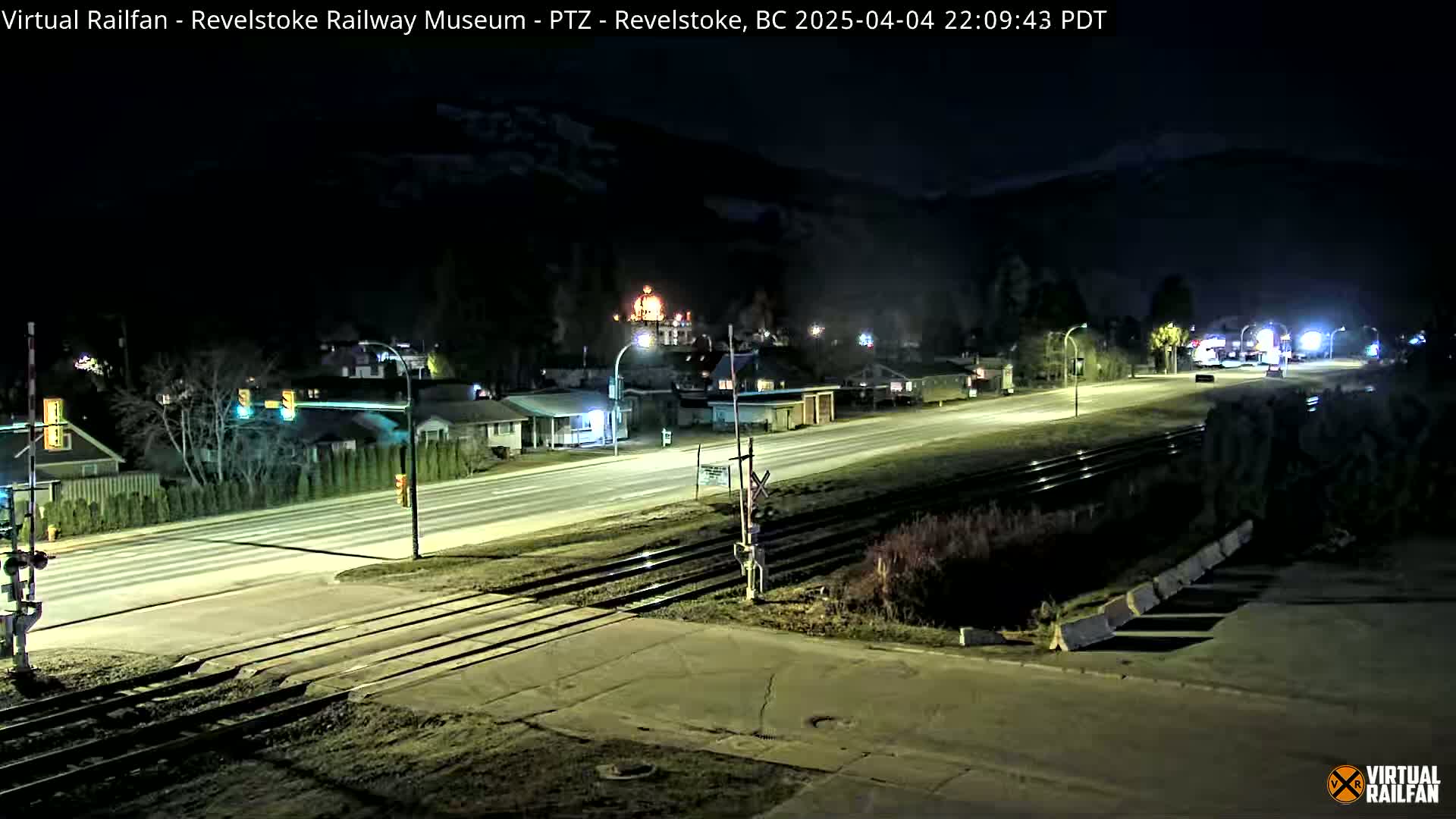 A nighttime view of a road and railway crossing in a small town, with dark mountains in the background under a clear sky.