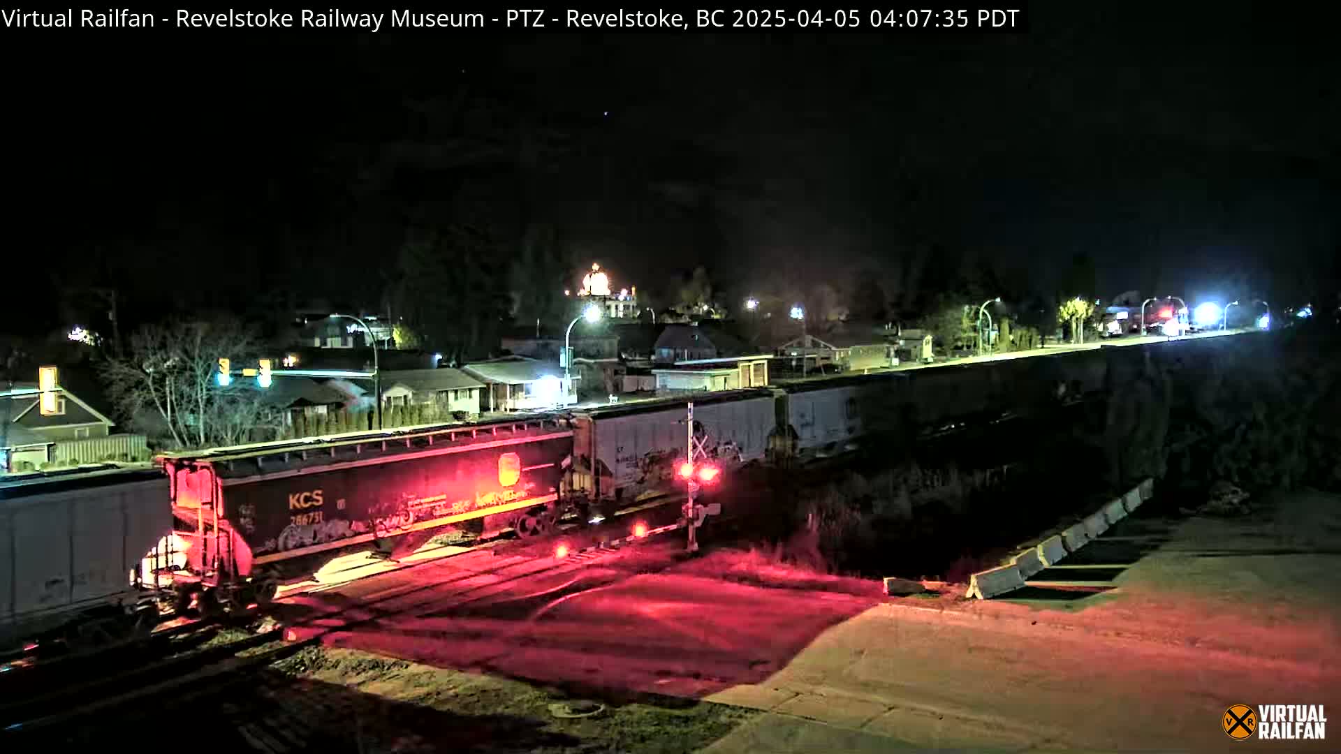 At night, a freight train with red lights illuminated passes a railroad crossing in a residential area.