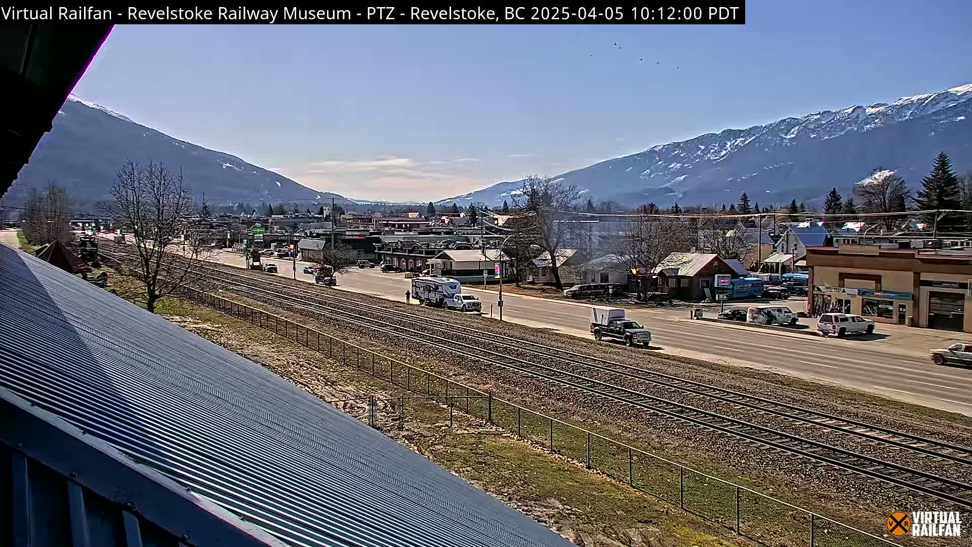 A sunny day reveals a view of a town nestled in a valley between snow-capped mountains, with a railway line running through it and vehicles traveling on a nearby road.