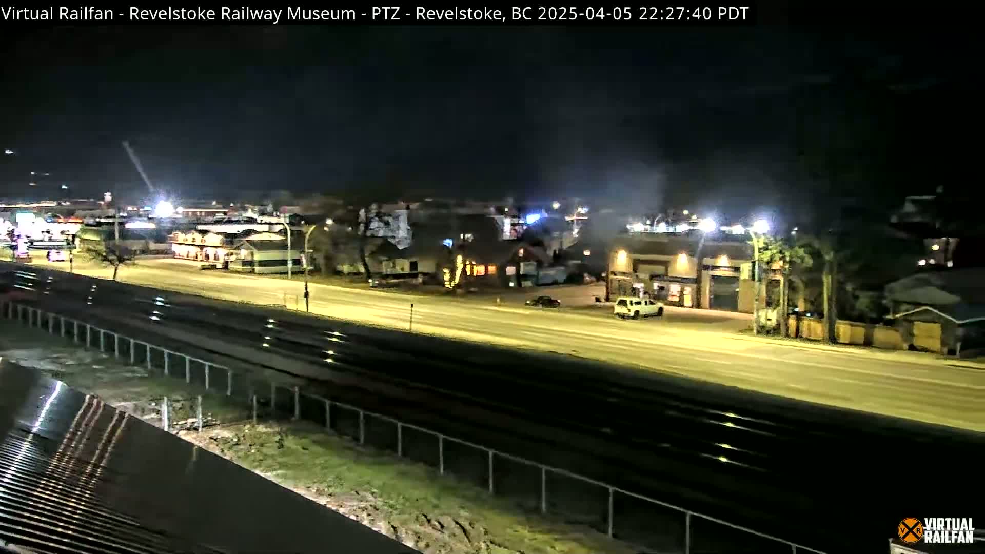 A nighttime view of a wet road and railroad tracks next to a sparsely populated area with buildings under lightly cloudy skies.