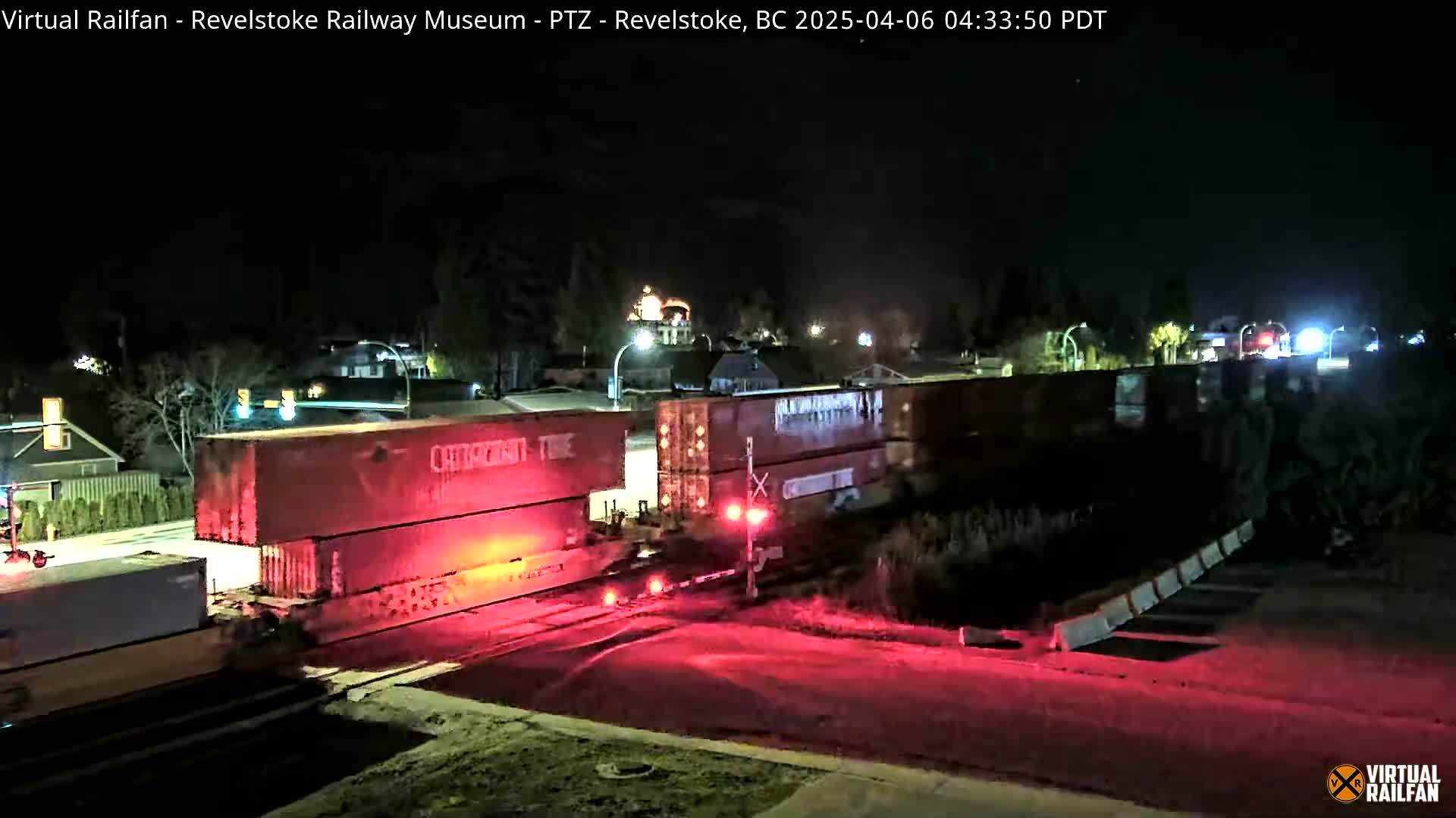 At night, a train with several red cargo containers is stopped at a railroad crossing, illuminated by red lights.