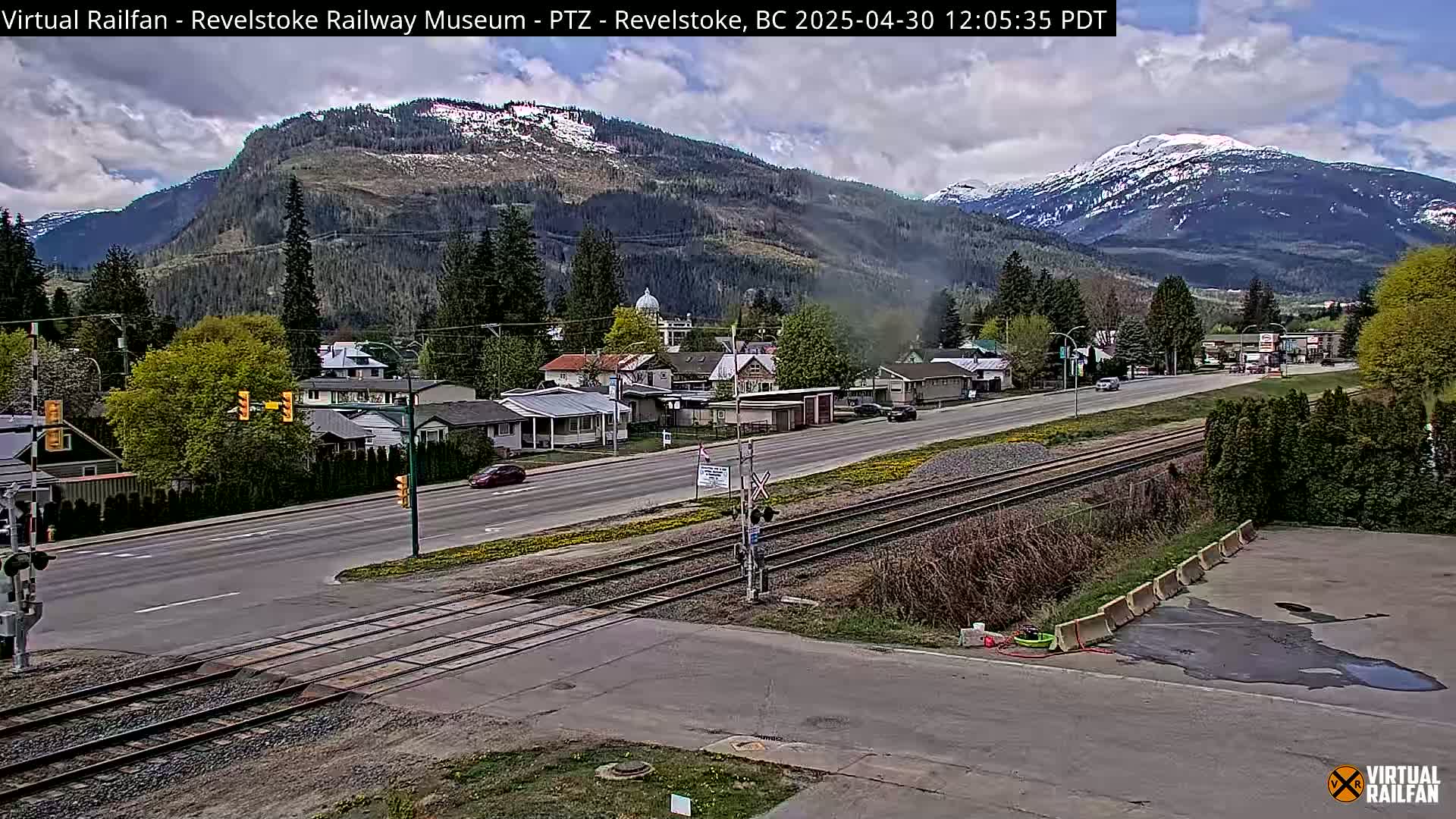 A partly cloudy day reveals a small town nestled in a valley, with snow-capped mountains in the background, and train tracks intersecting a roadway in the foreground.