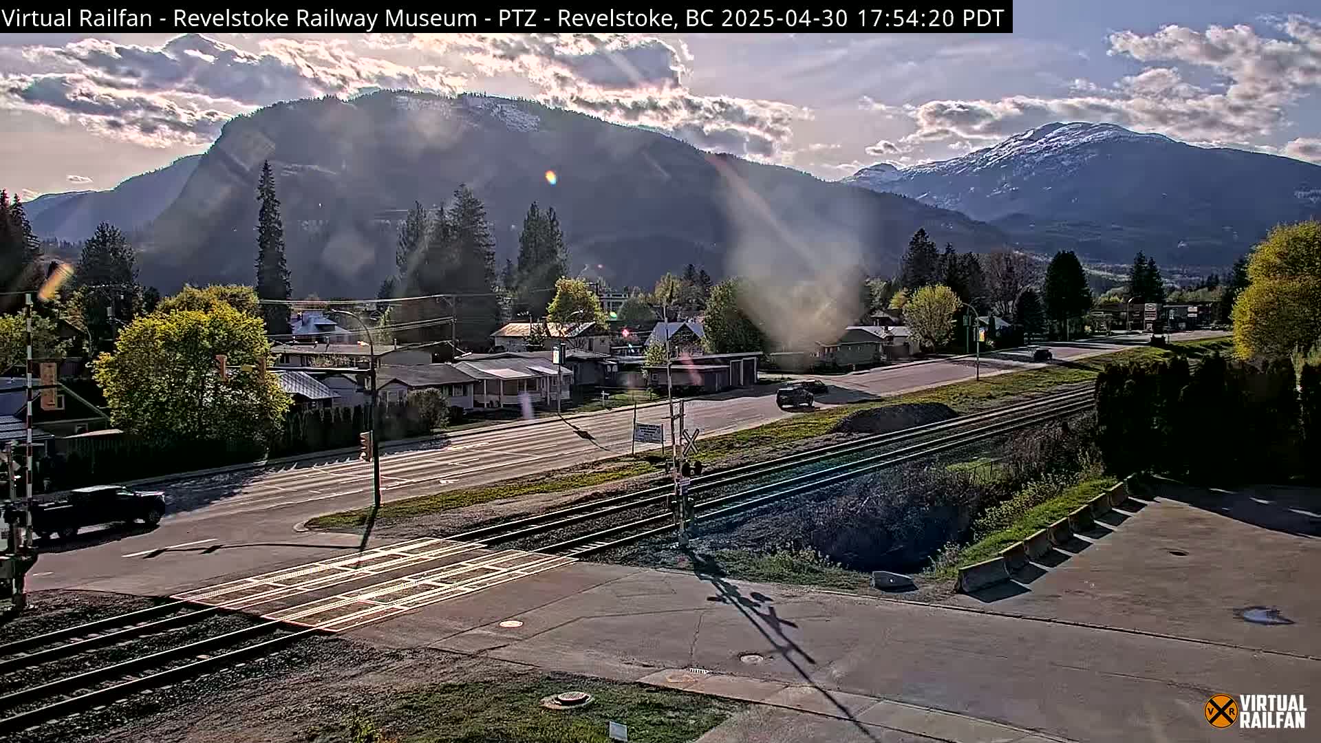 A partly sunny day reveals a mountain range behind a town with a railway crossing, residential homes, and sparse traffic.