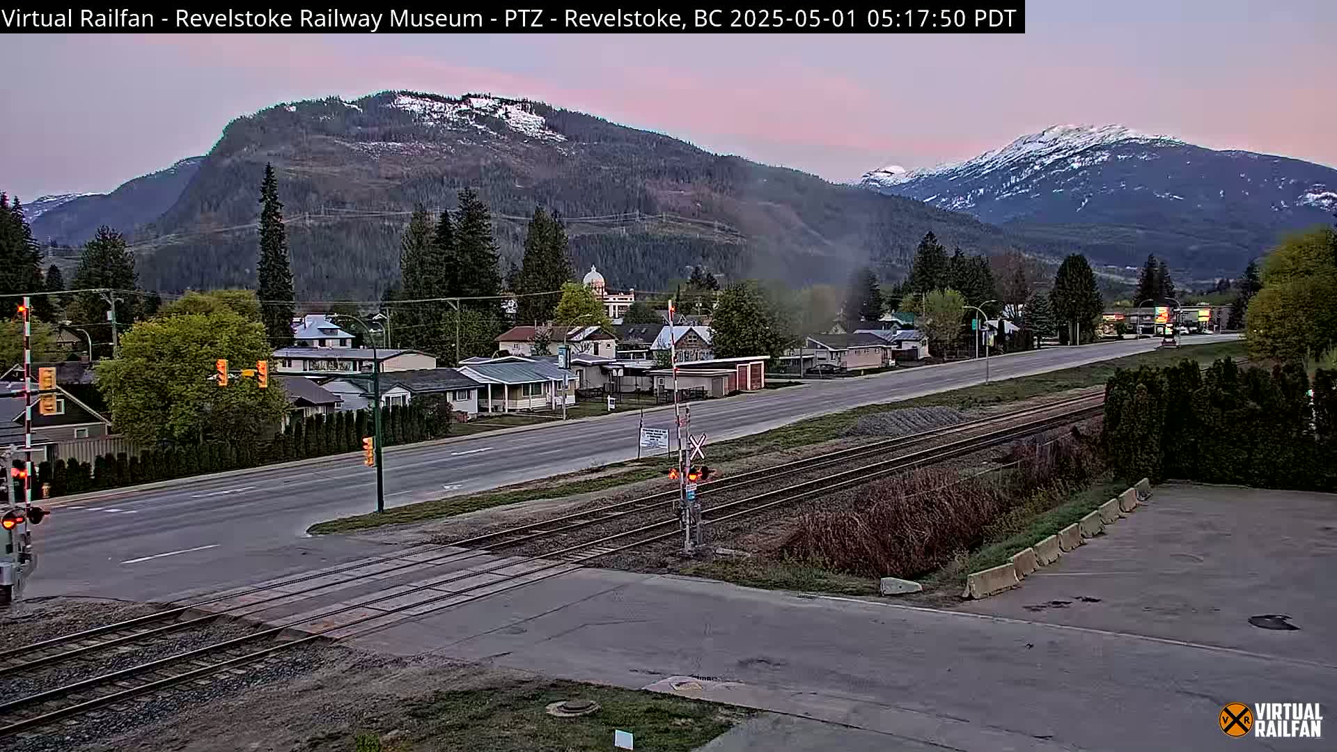 A view of a town nestled in a valley between snow-capped mountains at dawn, with a railway crossing in the foreground.