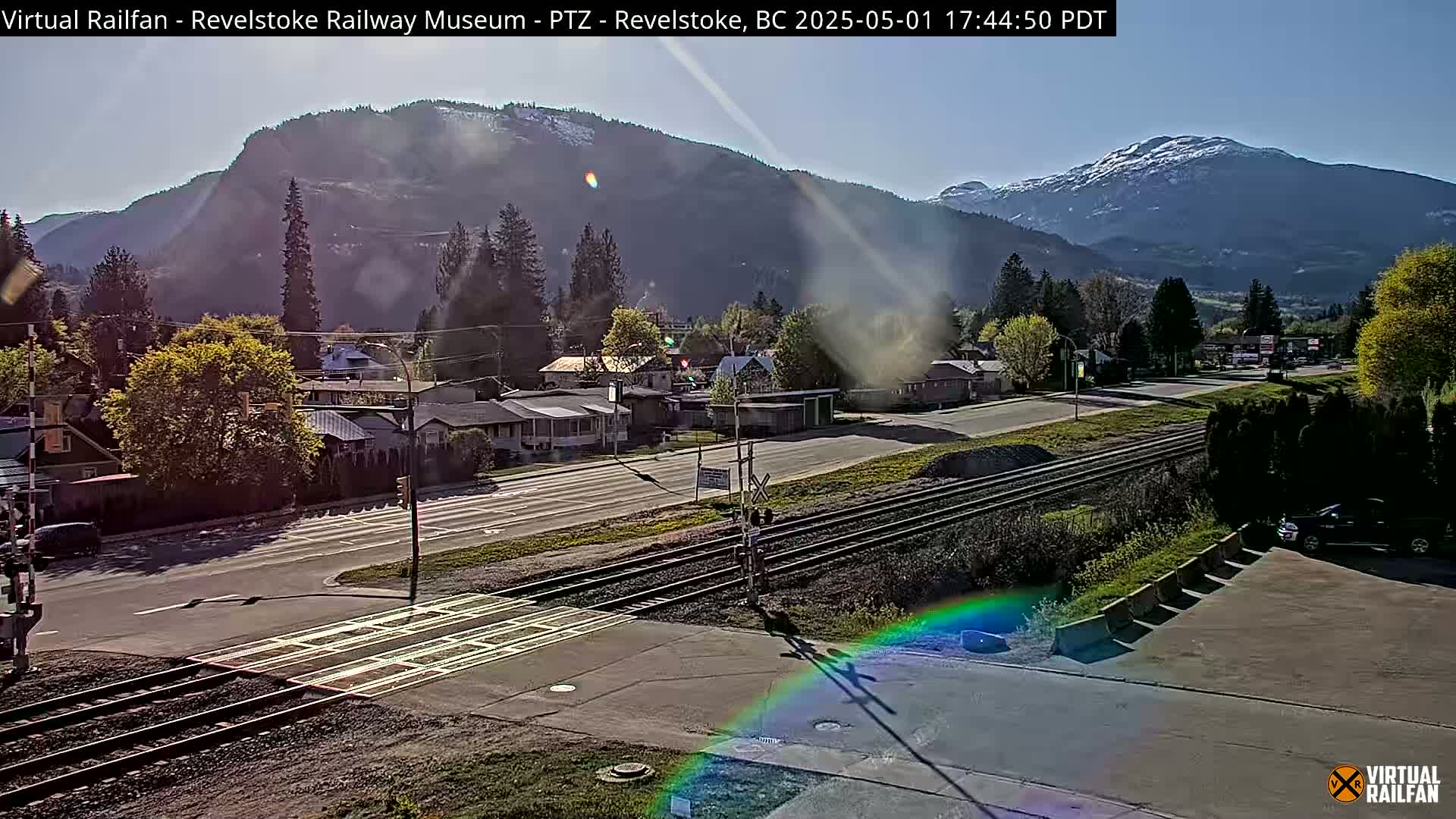 A sunny view of a town nestled in a valley between mountains, featuring a railroad crossing and a road.