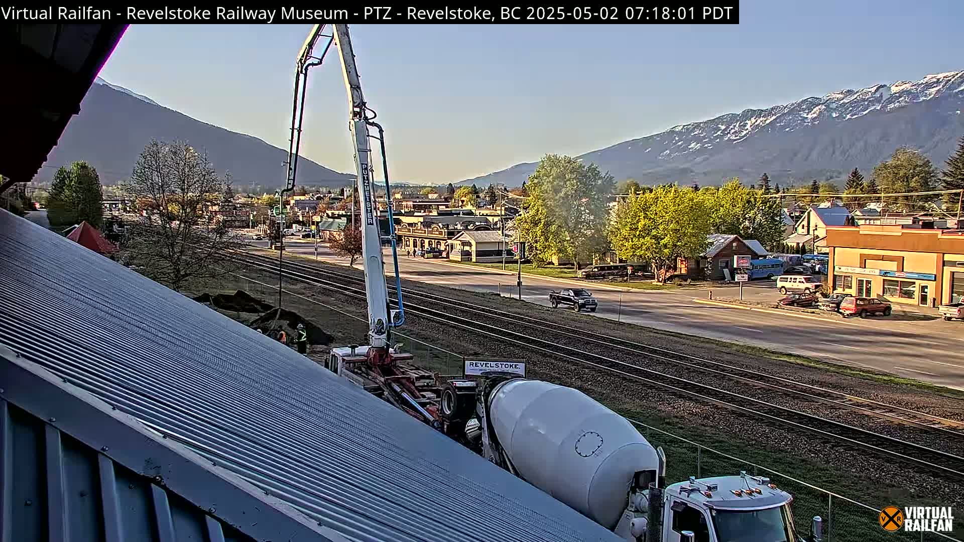 A cement mixer truck and a concrete pump are positioned on a sloped metal roof, with a town and mountains in the background under a clear sky.