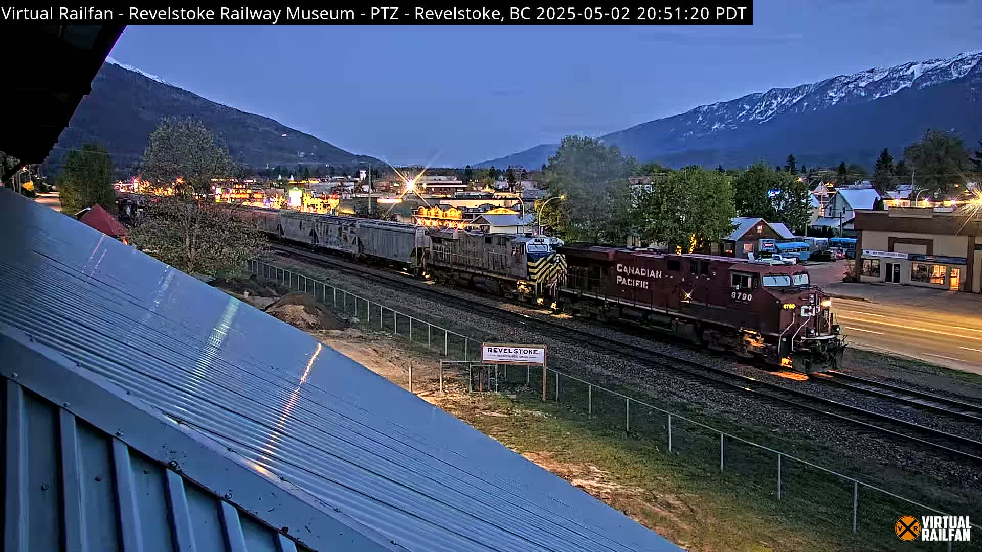 A long freight train passes through a small town at twilight, under a mostly clear sky with snow-capped mountains in the background.