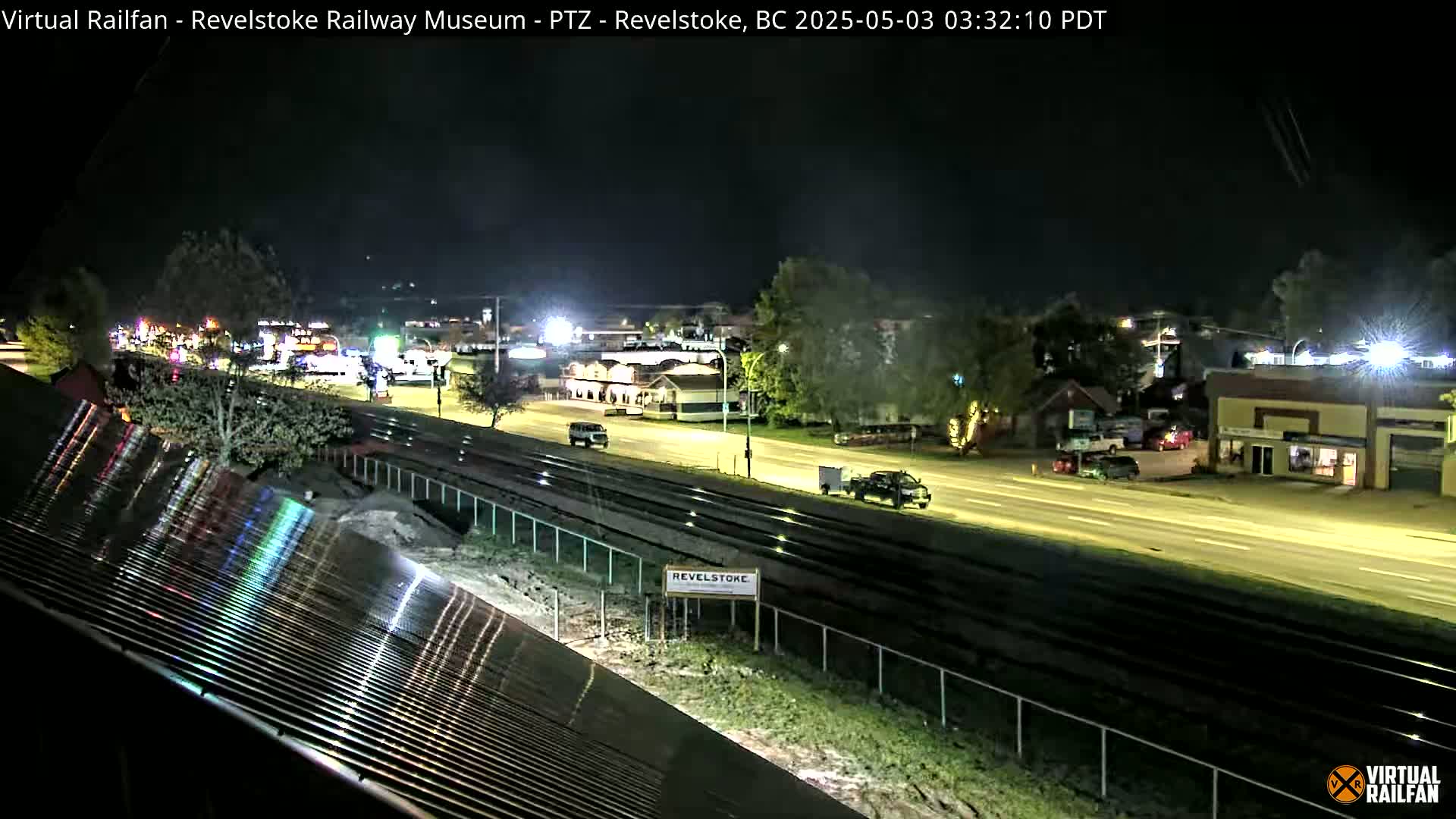 A nighttime view of a roadway with sparse traffic, adjacent to train tracks, and some buildings visible under partly cloudy skies.