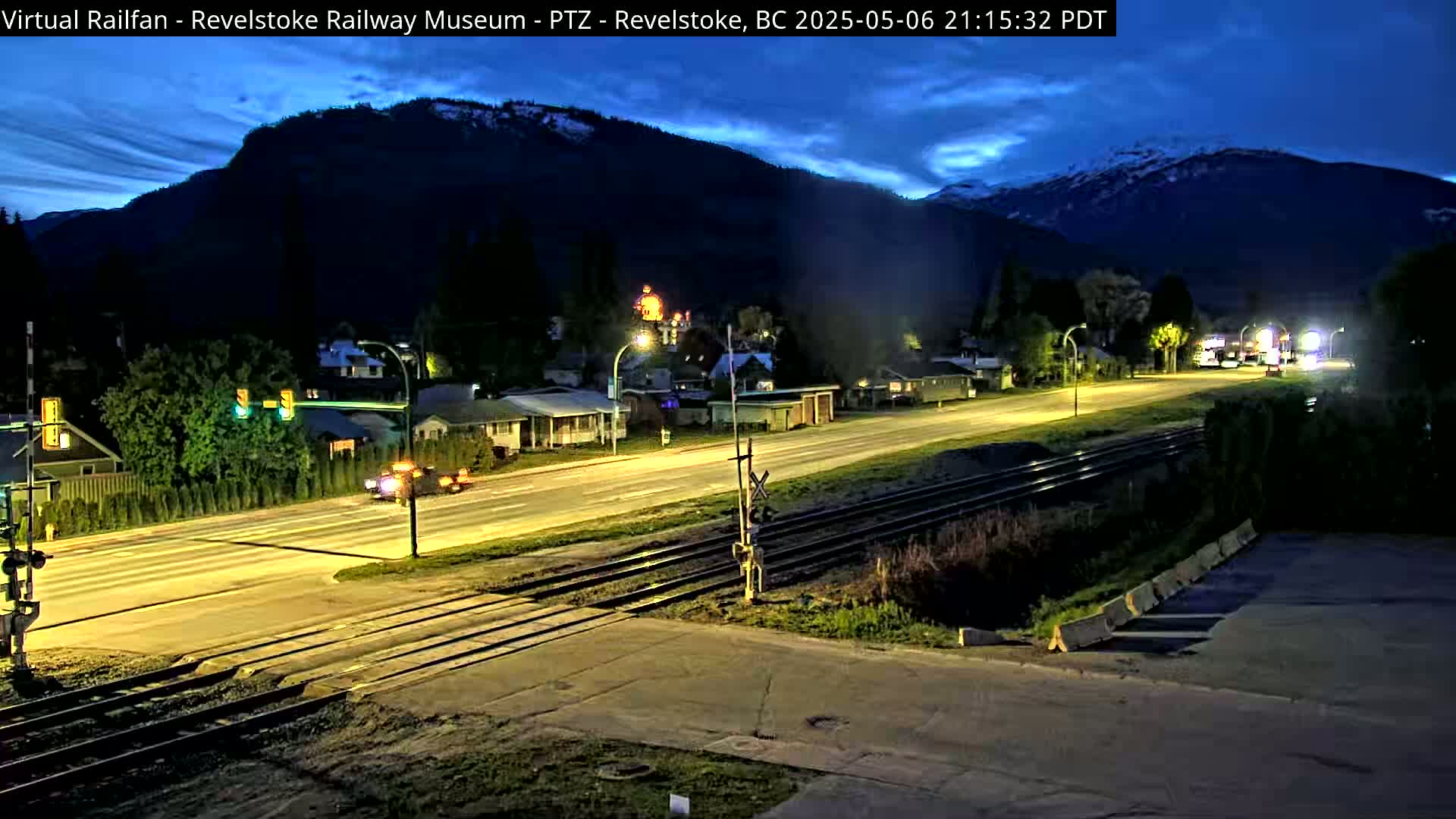 A nighttime view of a town under a twilight sky, showing a street with cars and train tracks running alongside it, all set against a backdrop of dark mountains.