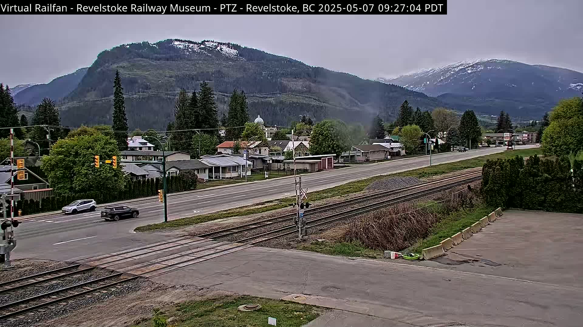 A partly cloudy day shows a view of a town with a railroad track intersecting a road, nestled between mountains with some snow on the peaks.