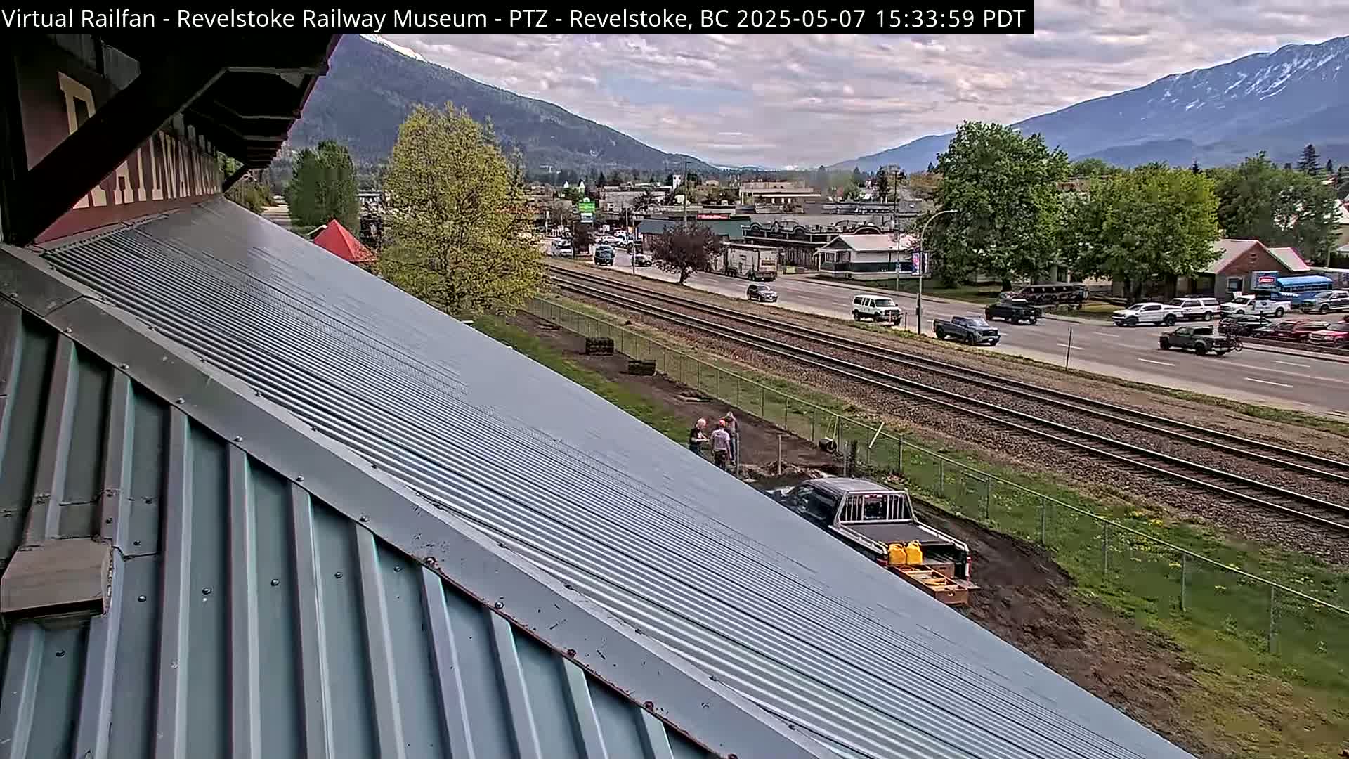 A mostly cloudy day shows a corrugated metal roof angled down towards a street scene with railway tracks, vehicles, and a few people working near a truck.
