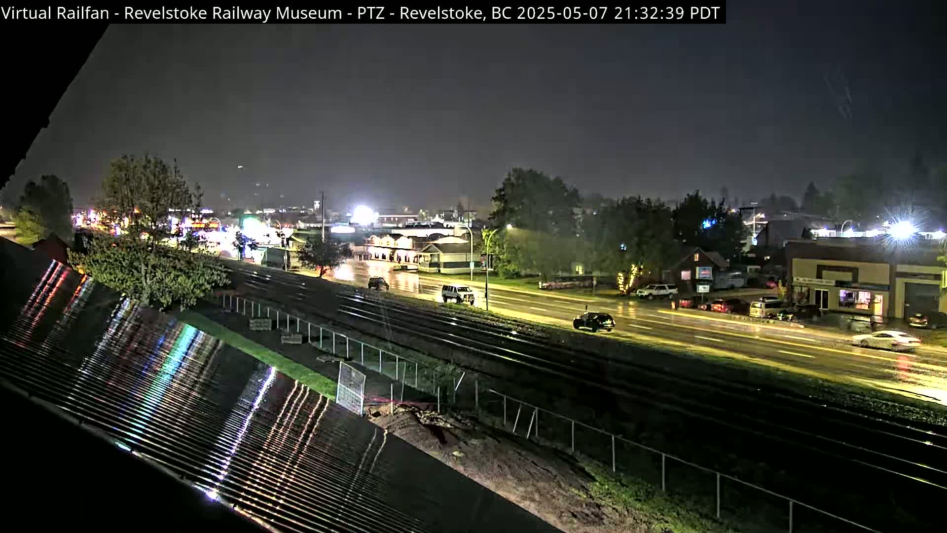 A nighttime, rainy view of a highway and train tracks near buildings and trees.