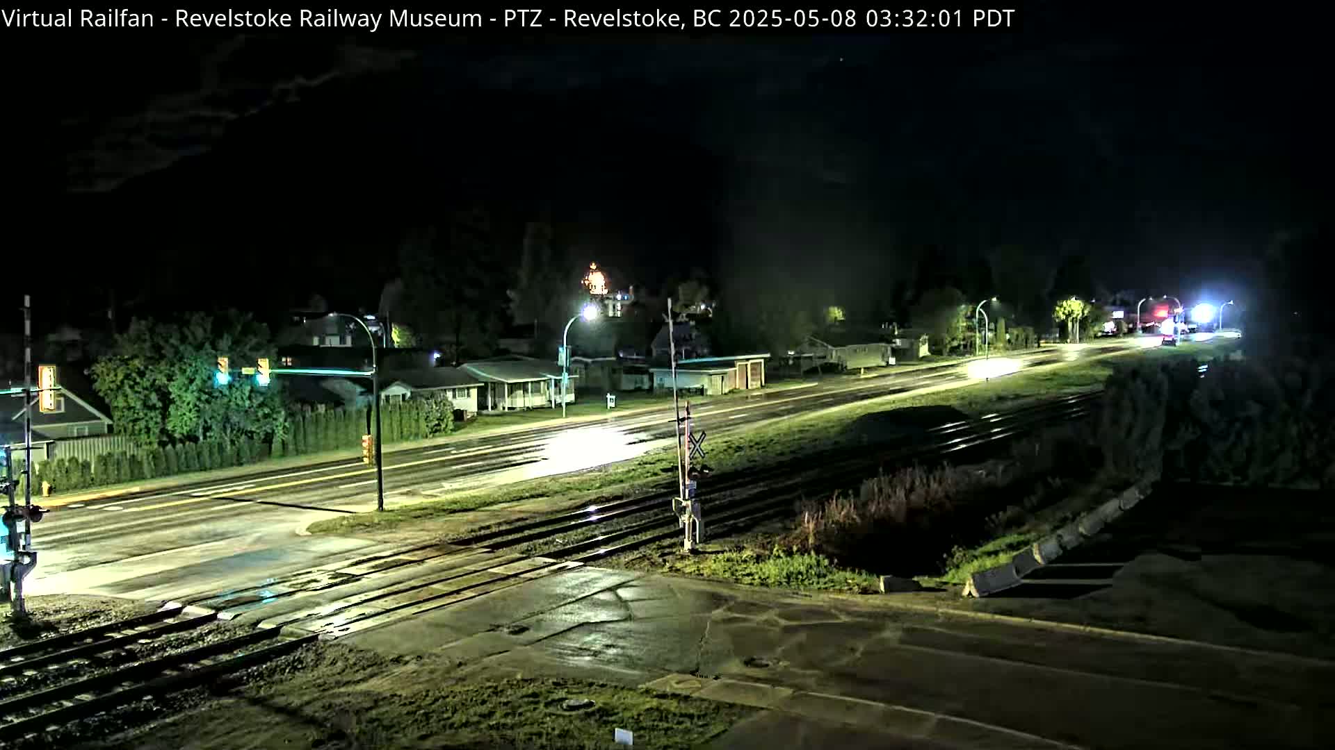 A nighttime view of a wet street and railroad crossing, with sparsely lit houses and trees in the background under a partly cloudy sky.