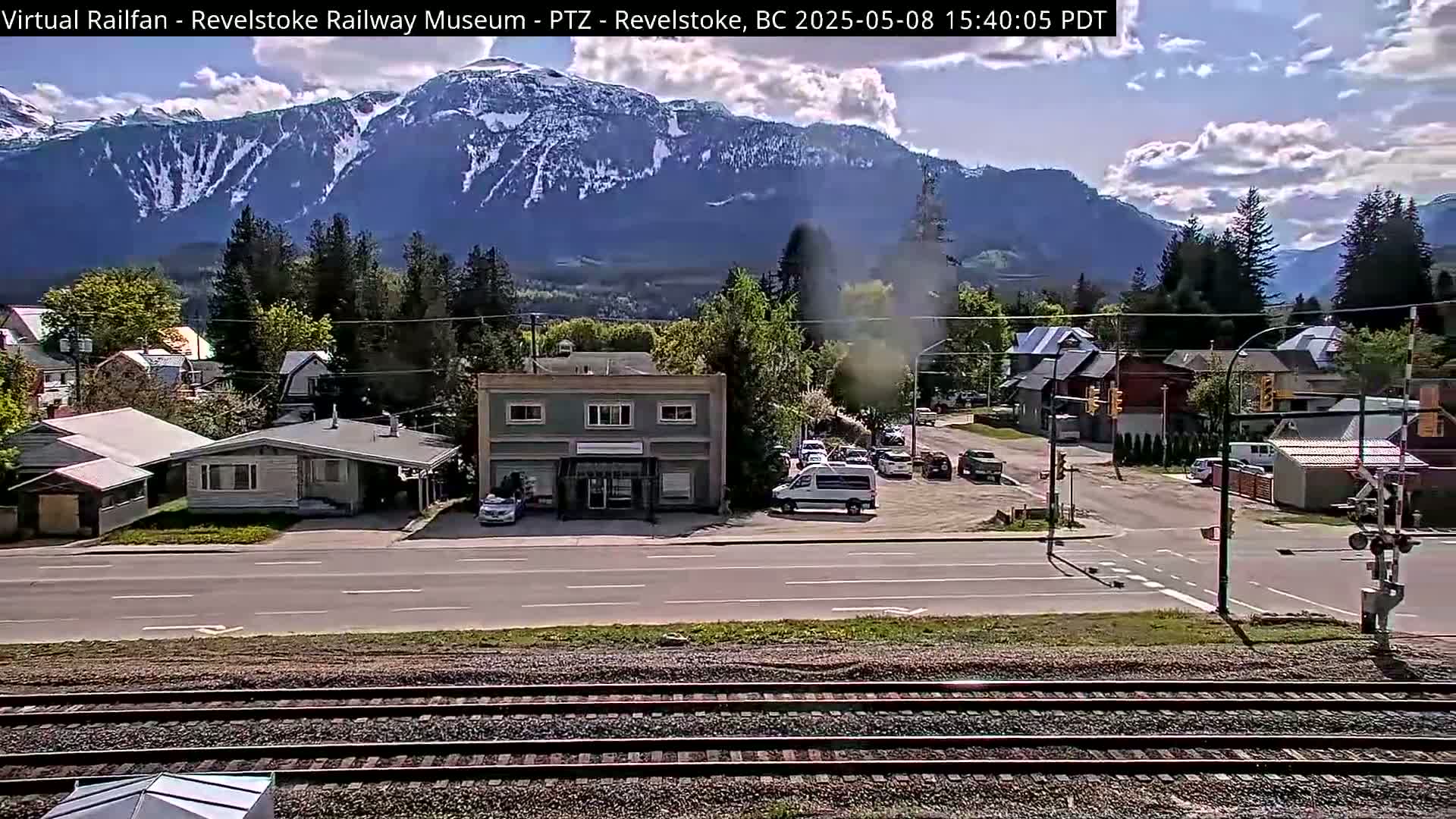 A partly cloudy day shows a mountain range in the background, overlooking a town with buildings, vehicles, and railroad tracks in the foreground.