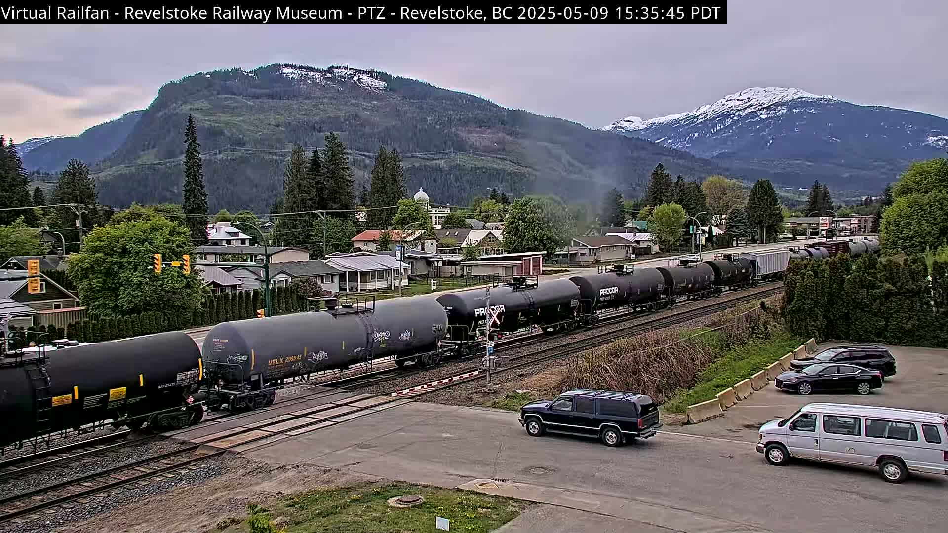A long train of tanker cars passes through a residential area nestled against a mountain range partially covered in snow under a cloudy sky.