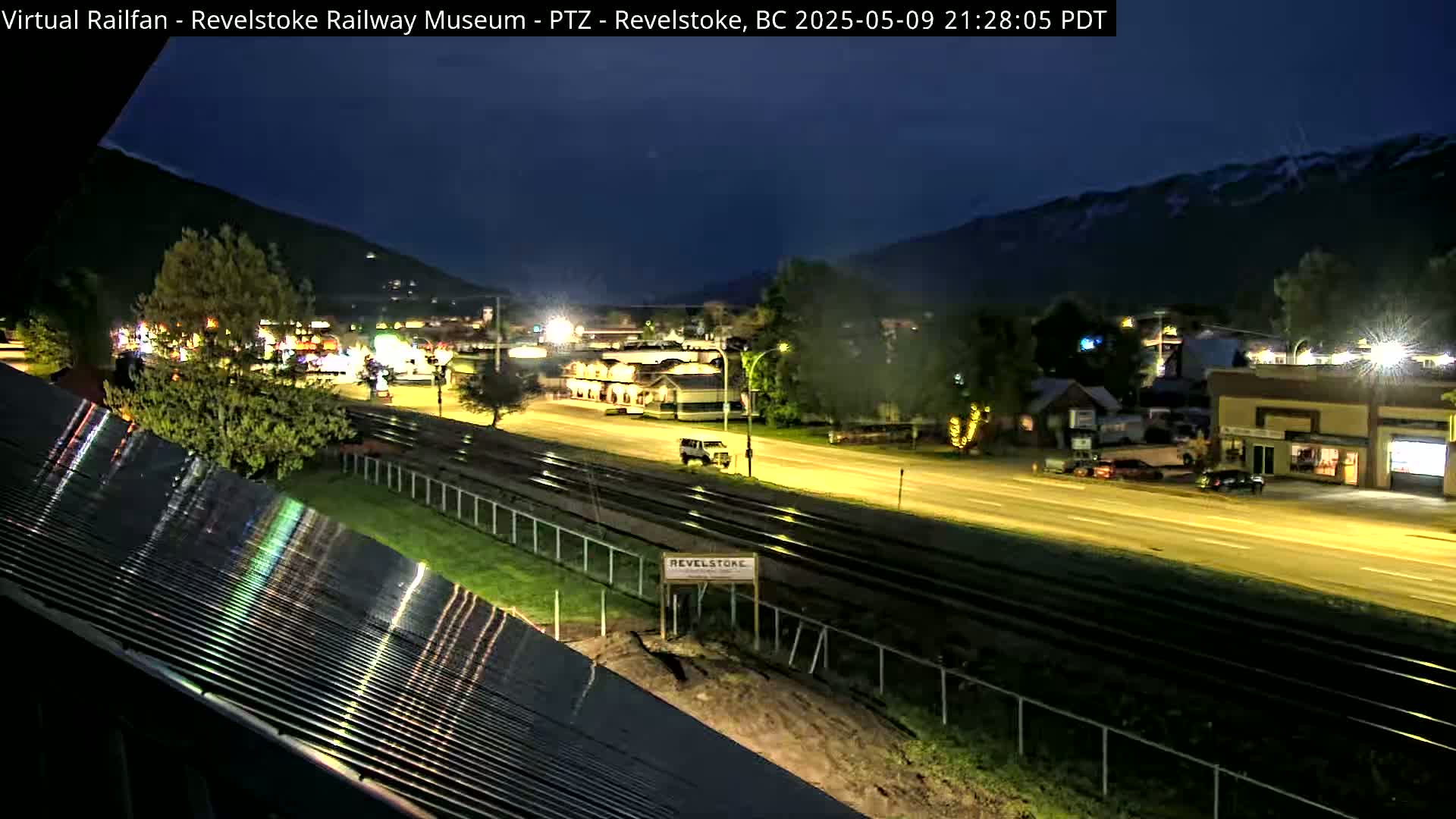 A nighttime view of a town situated beside a mountain range, illuminated by streetlights, with train tracks running parallel to a road.