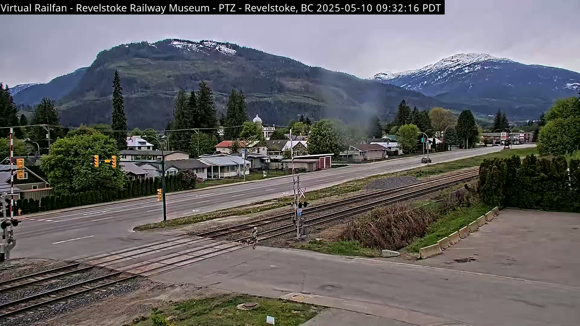 A partly cloudy day reveals a town nestled at the base of snow-capped mountains, with a railroad crossing in the foreground.