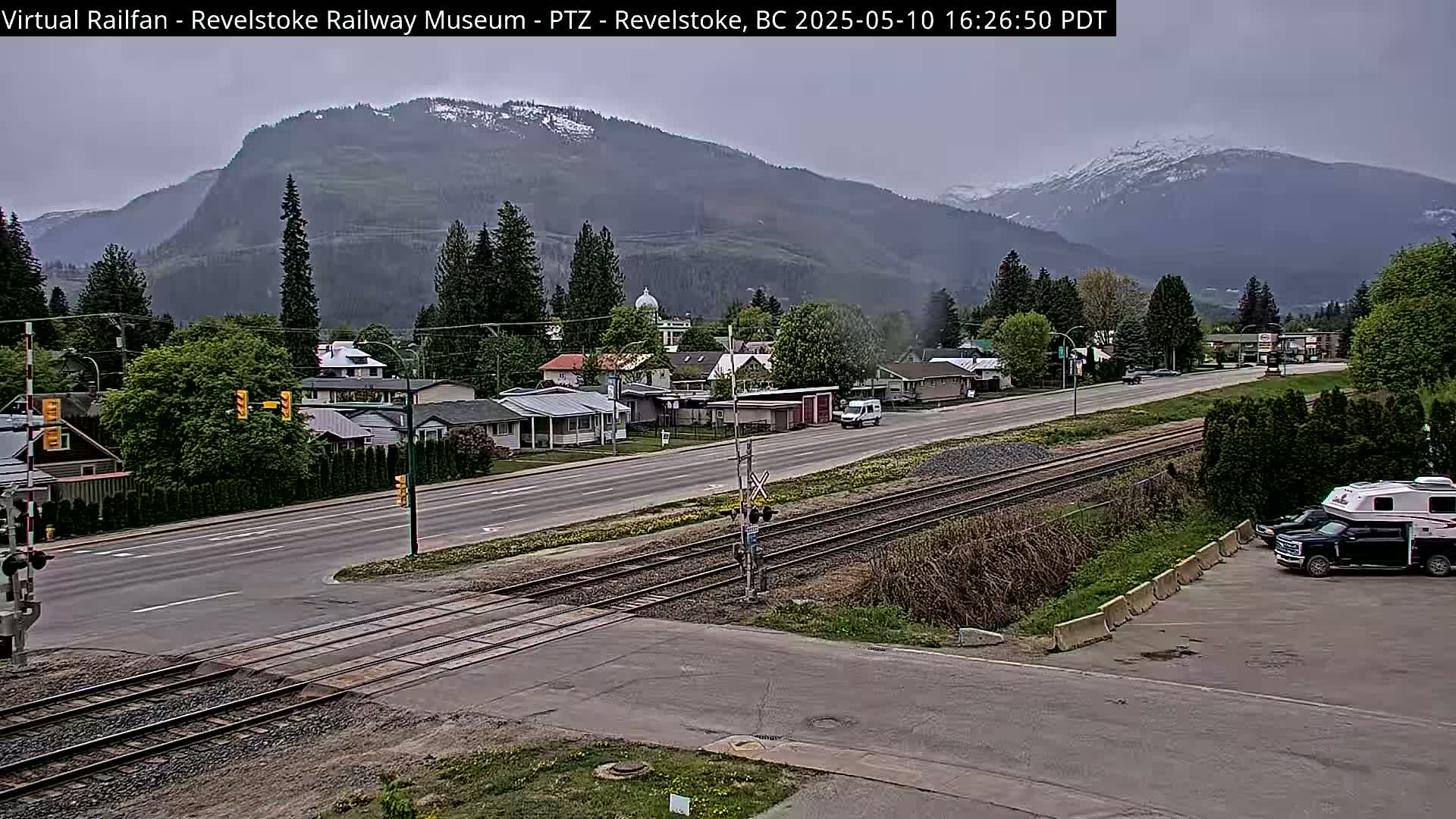 A mostly cloudy day shows a town nestled in a valley with mountains in the background, railroad tracks crossing a road in the foreground.