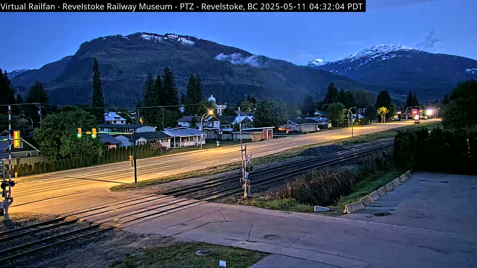 A twilight view of a town nestled in a valley, featuring a highway crossing train tracks, with mountains in the background showing some snow.