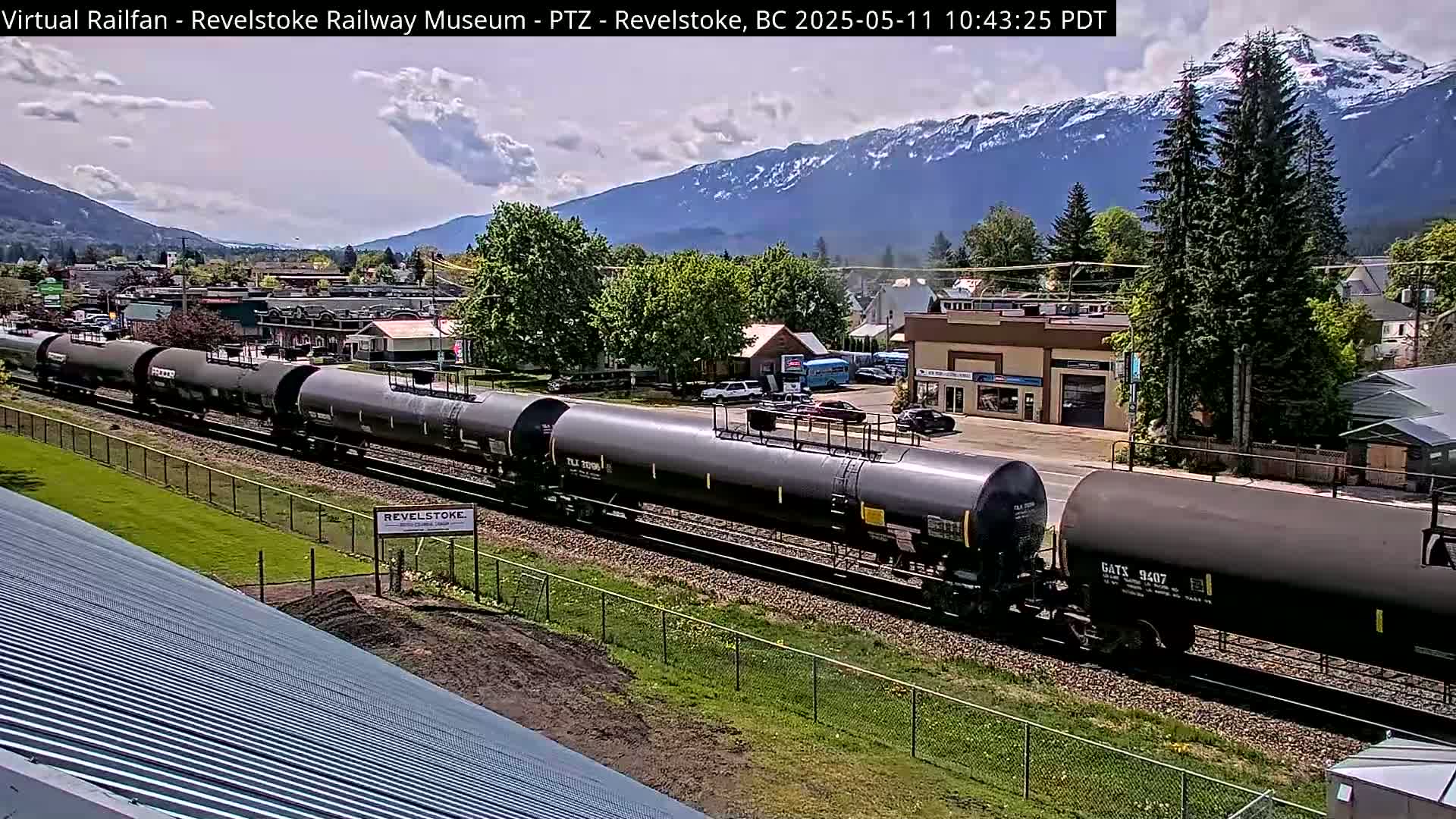 A long train of dark tanker cars passes through a small town nestled in a valley, with snow-capped mountains visible in the background under a partly cloudy sky.