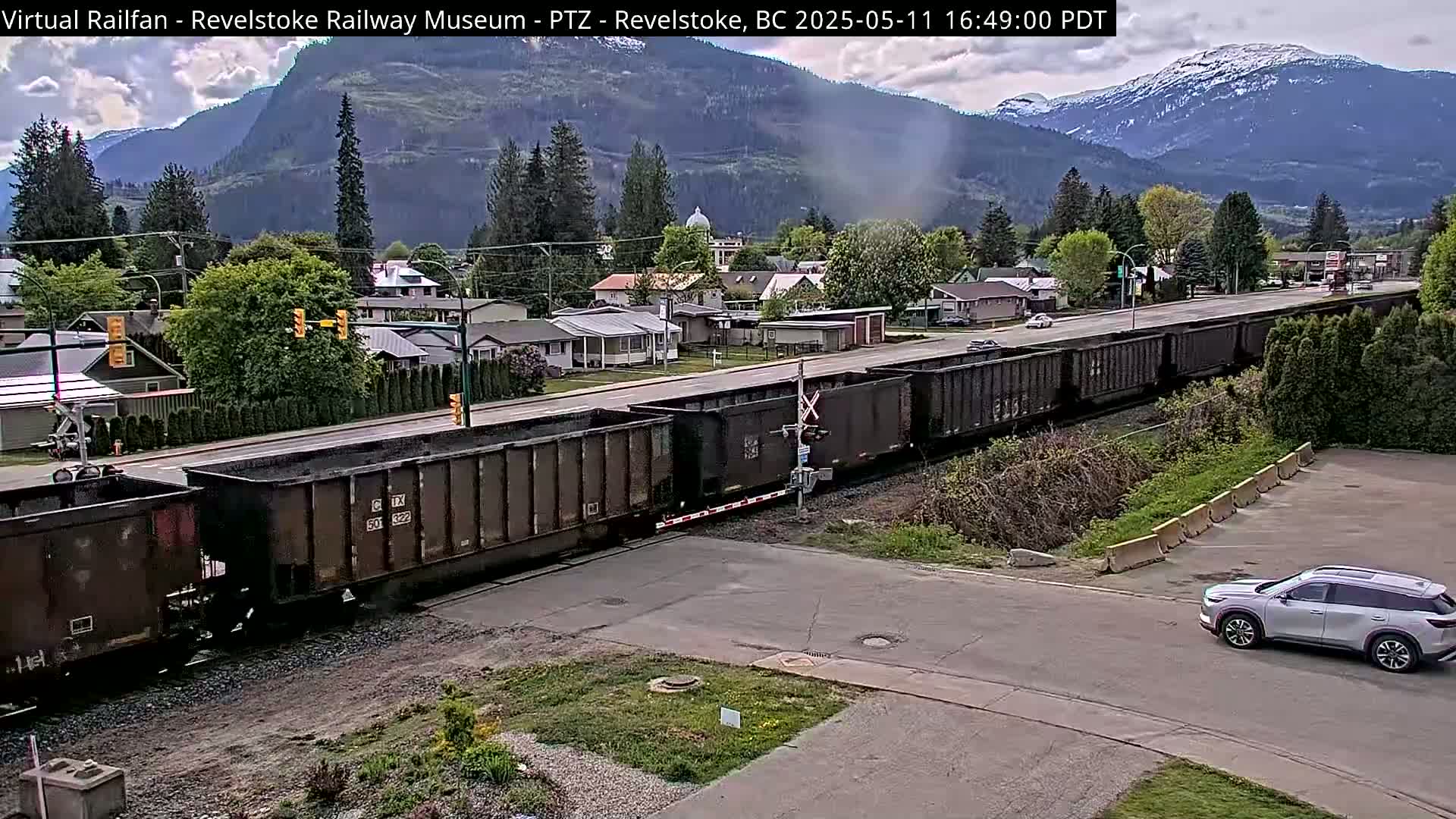 A long freight train crosses a road in front of a small town nestled in a valley, with mountains in the background under partly cloudy skies.