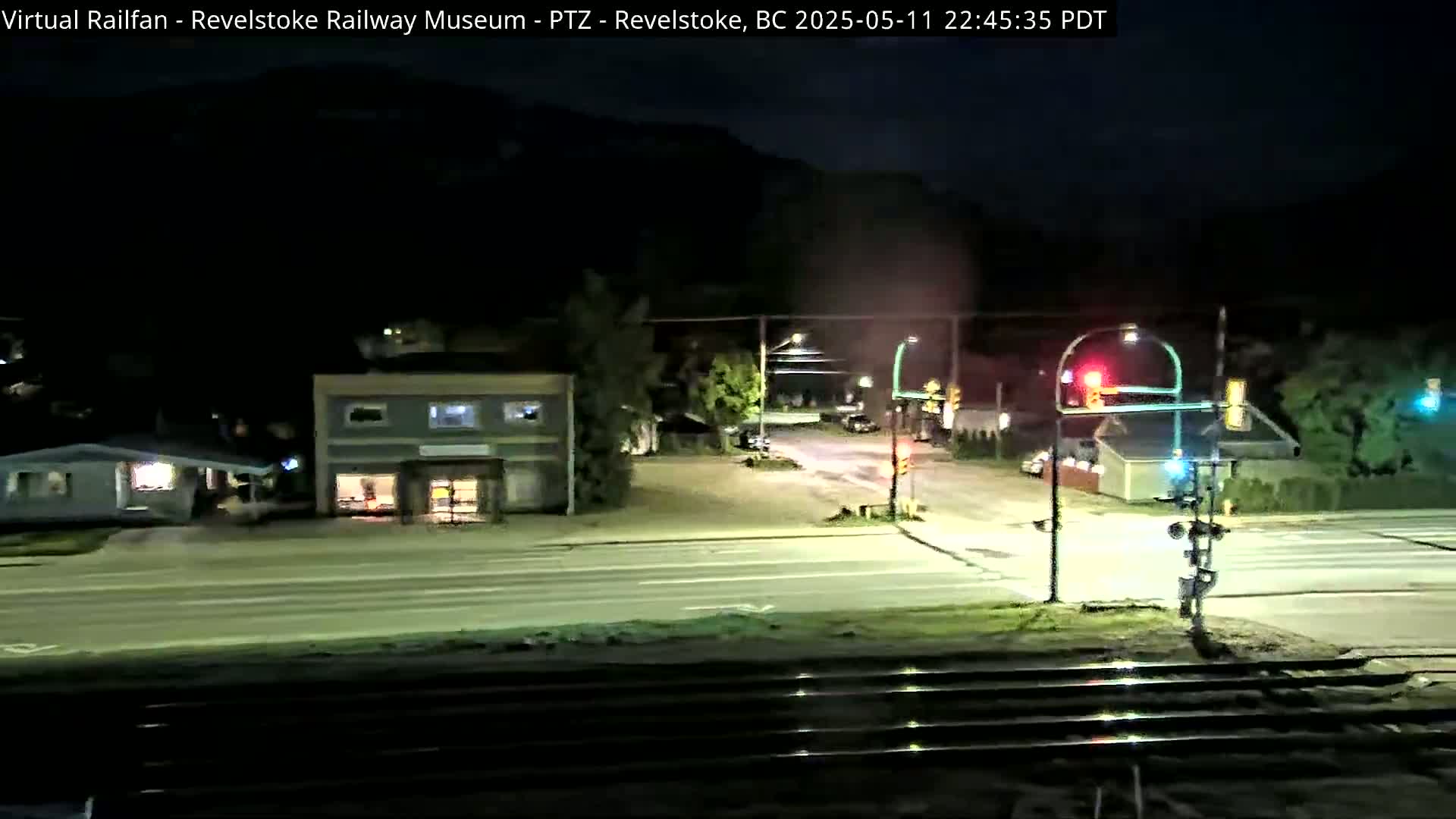 At night, a mostly empty street intersection with a few buildings and train tracks is viewed from above.