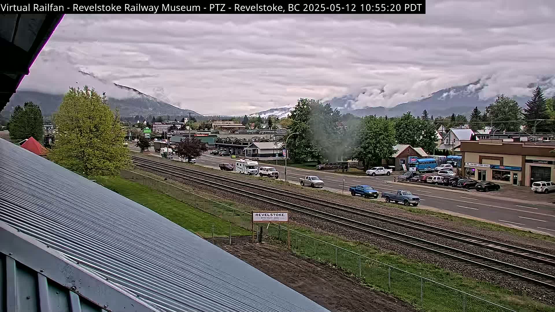 A view of a town with railroad tracks and vehicles, under a cloudy sky, with mountains partially obscured by clouds in the background.