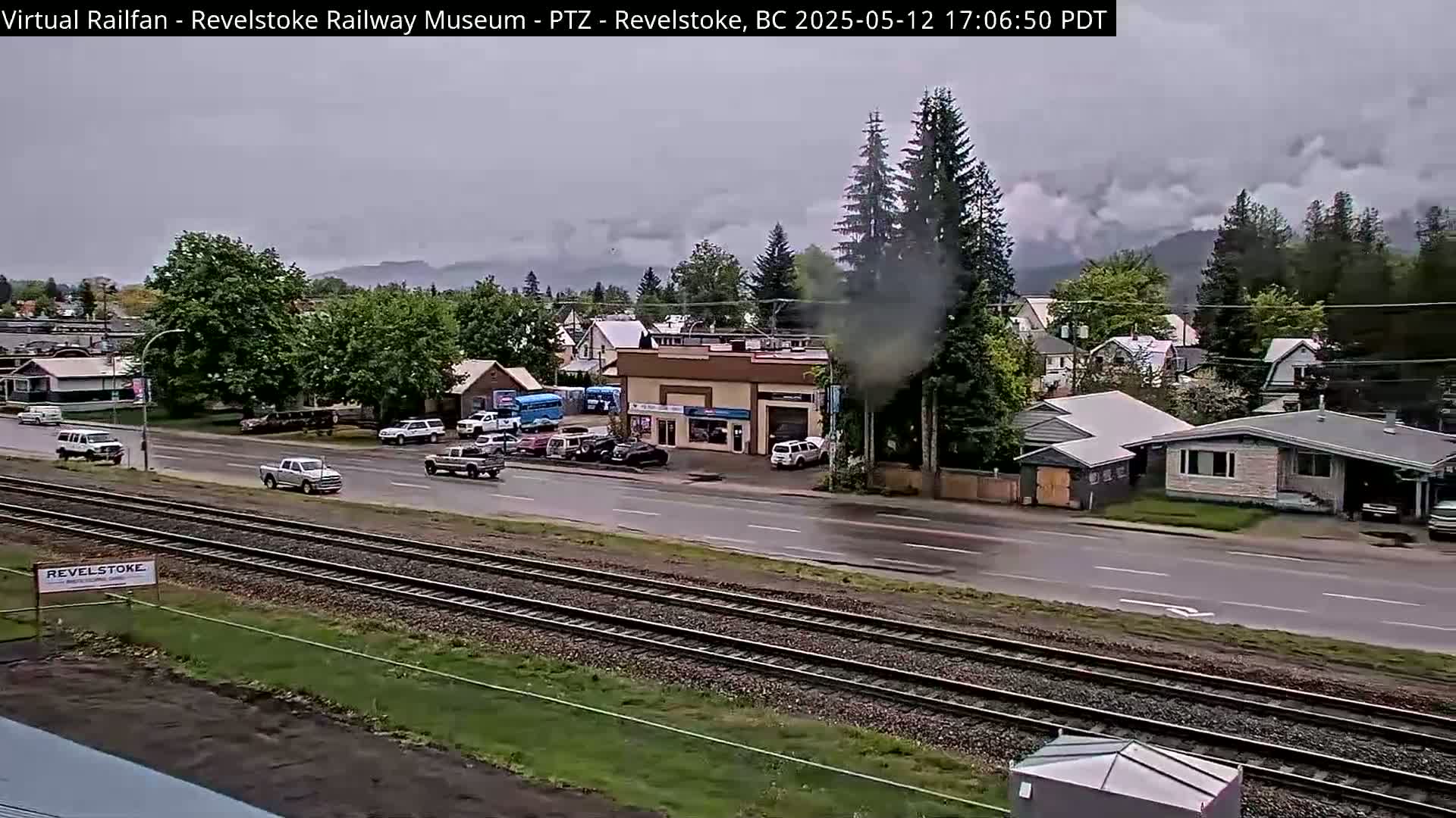 A view of a town under an overcast sky, showing several vehicles on a road that runs parallel to a set of train tracks.
