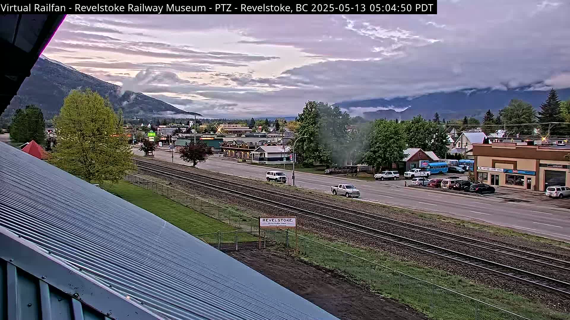 A partly cloudy sunrise illuminates a town with a railway track running through it, nestled between mountains shrouded in mist.