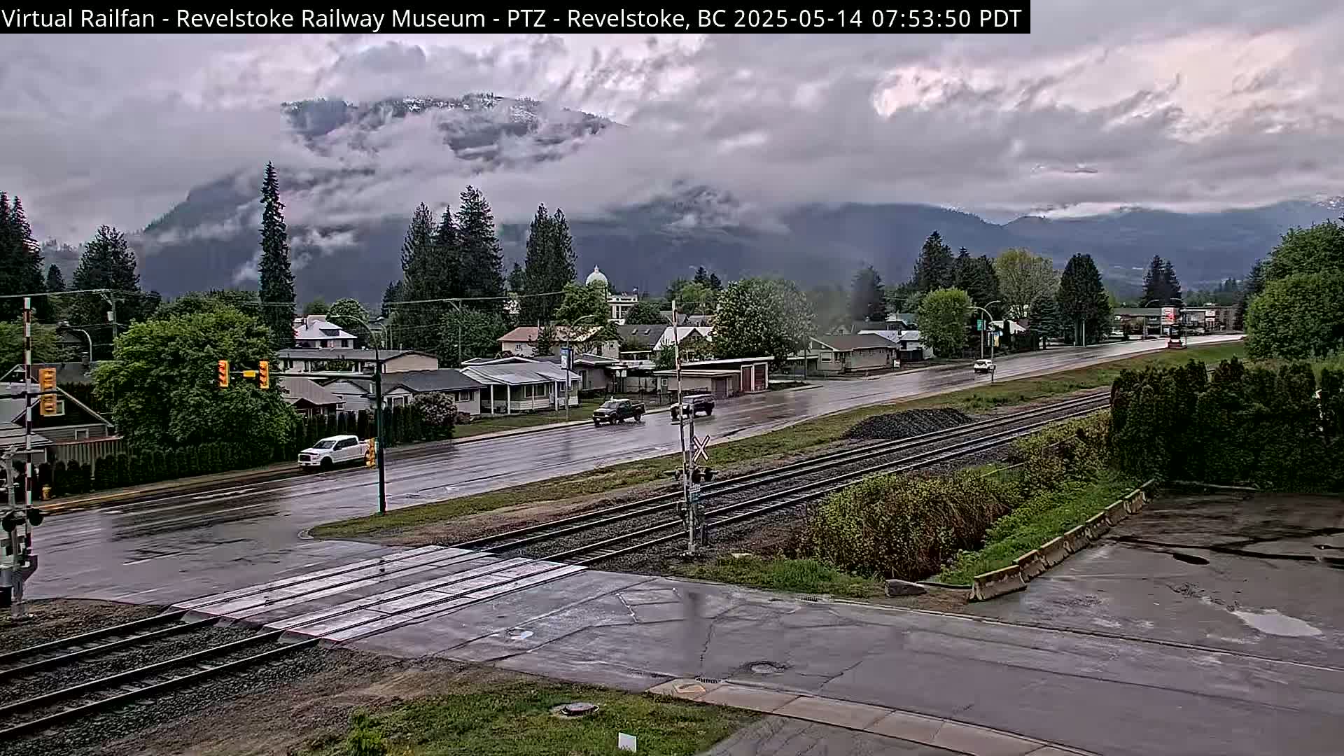 A wet town with a railroad crossing is nestled in a valley beneath overcast mountains.