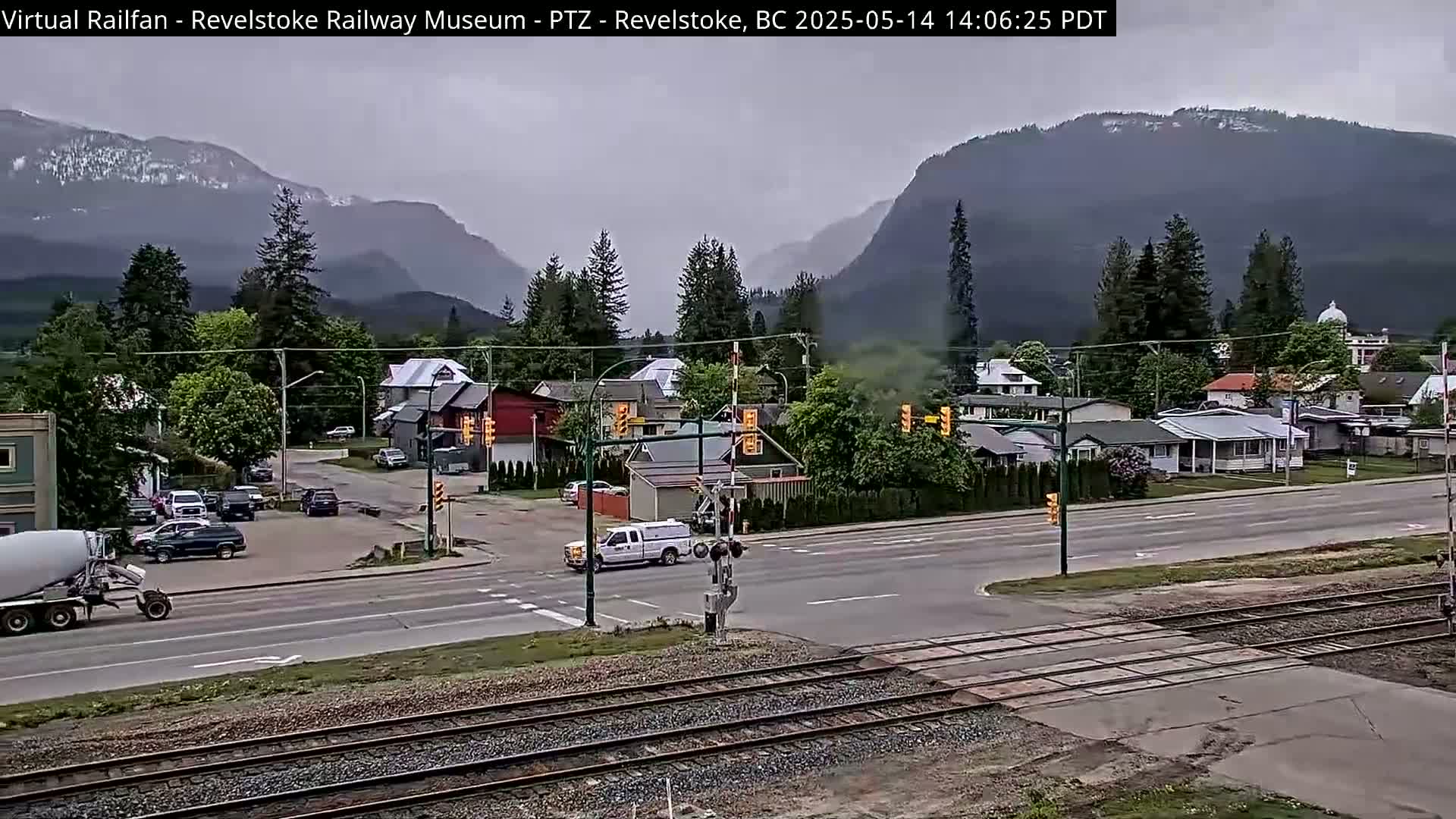 A cement mixer truck drives past a railway crossing in a town nestled at the foot of overcast mountains.
