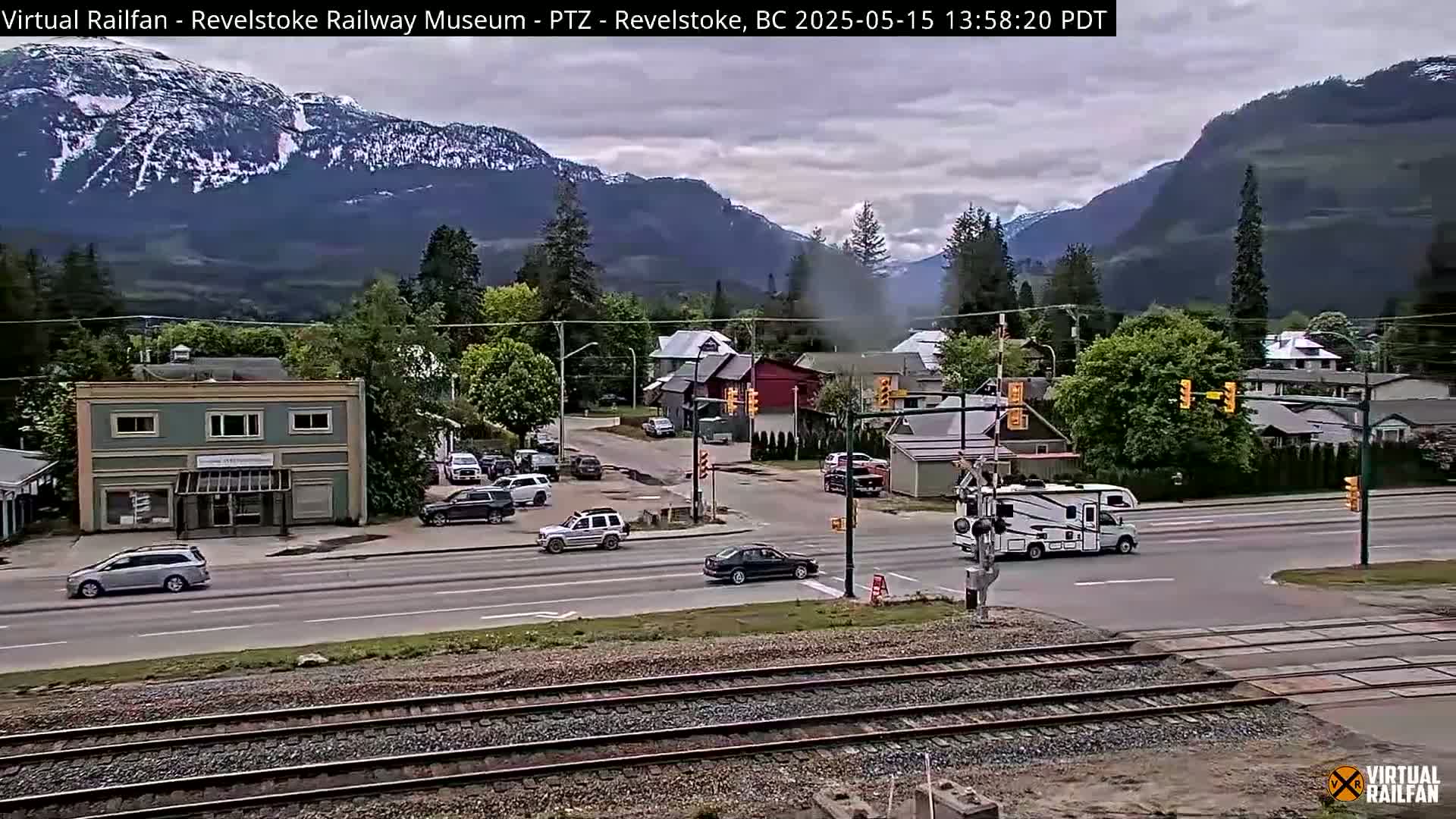 A cloudy day in a mountain town shows several vehicles at an intersection near train tracks, with a mix of residential and commercial buildings.
