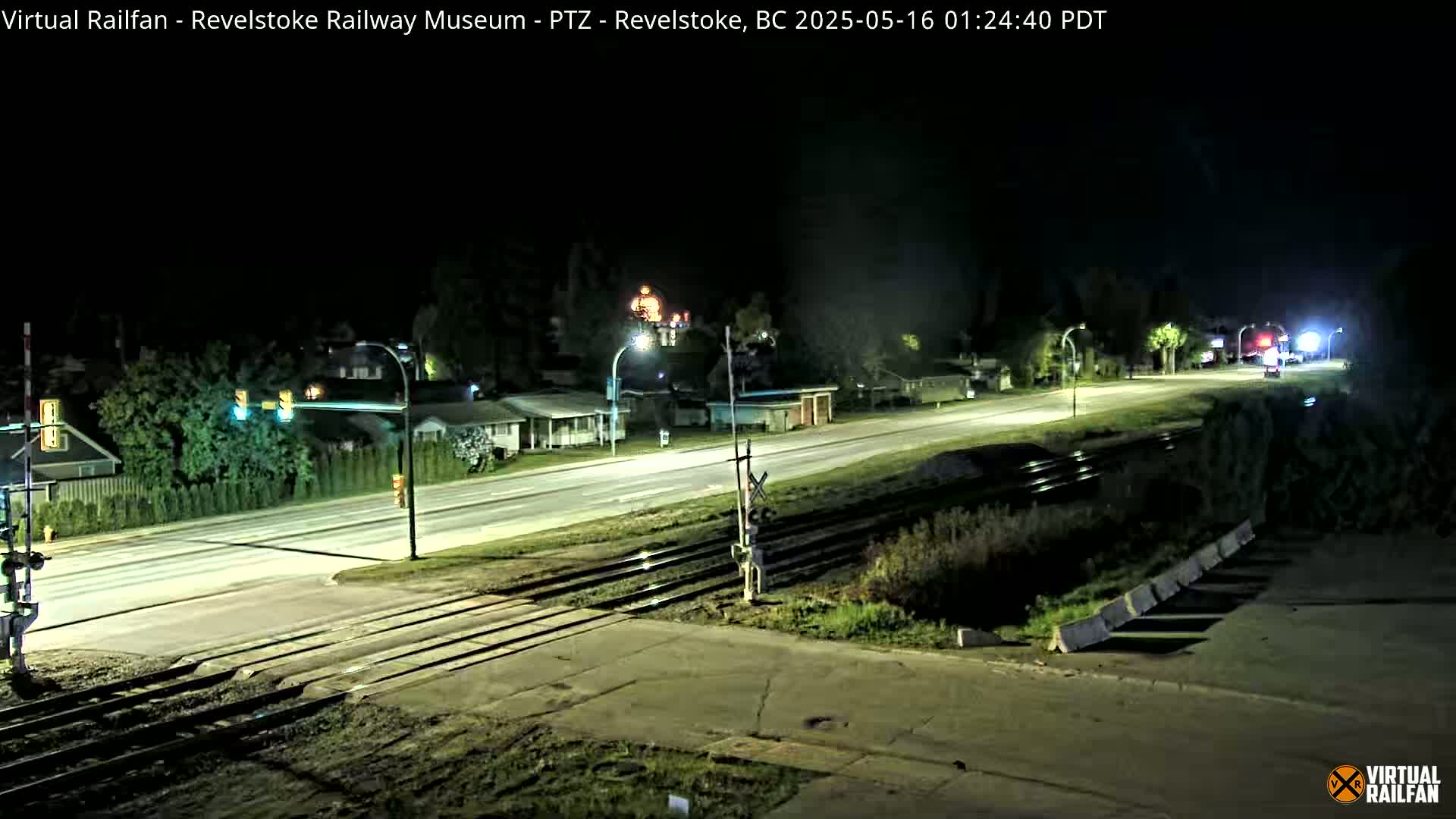 A nighttime view of a road crossing railroad tracks, with houses and streetlights visible in the background under a clear, dark sky.