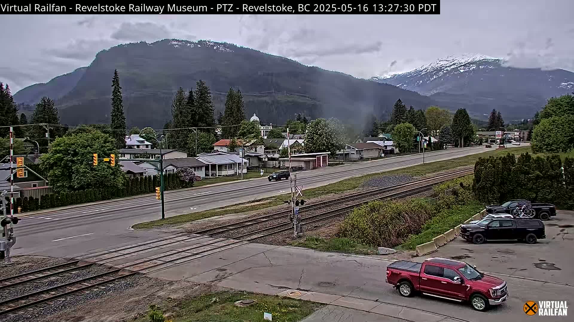 A partly cloudy day reveals a mountain range with snow-capped peaks overlooking a town with a highway, railway tracks, and several vehicles.