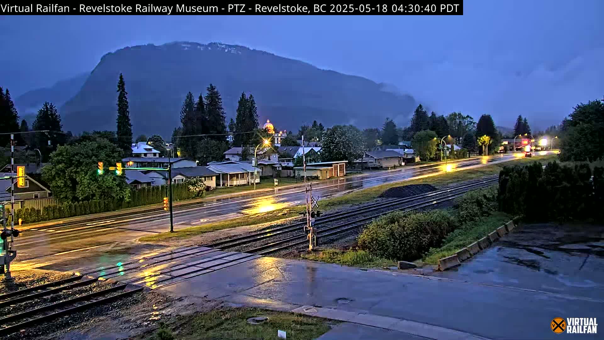 A rain-slicked town at twilight, with a mountain looming in the background, is intersected by railway tracks.