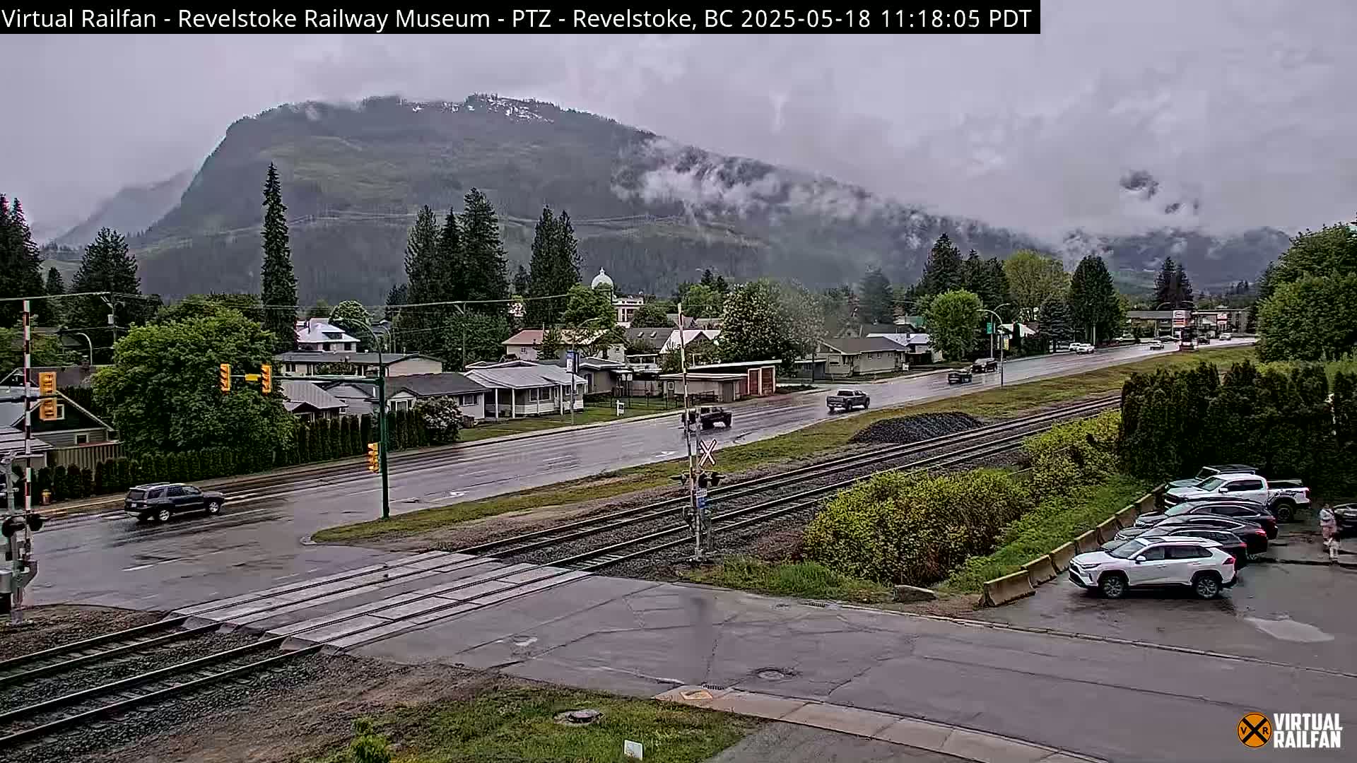 A wet, overcast town scene shows a road intersecting with train tracks, a few cars on the road and parked, and a mountain range partially obscured by clouds in the background.