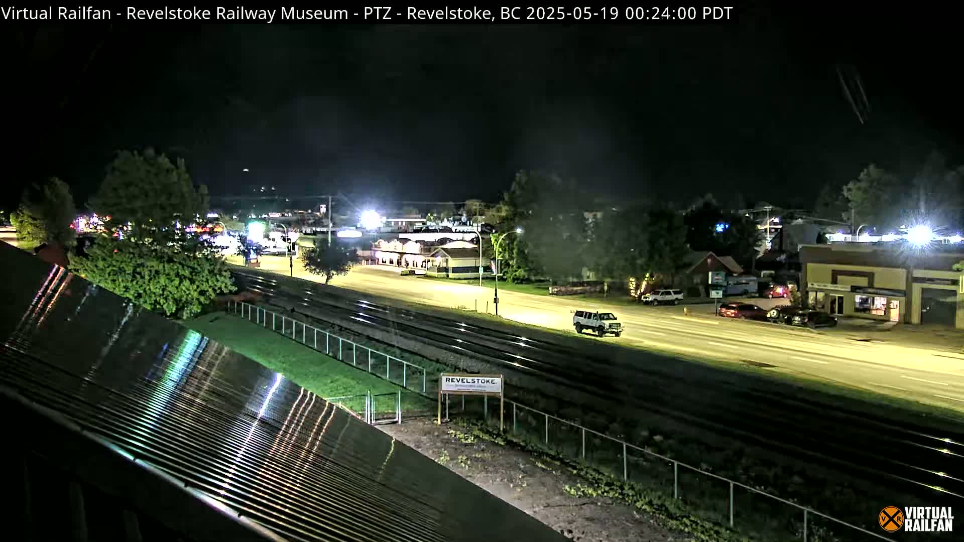 A nighttime view of a roadway and train tracks, with a few vehicles visible and trees lining the sides of the road.