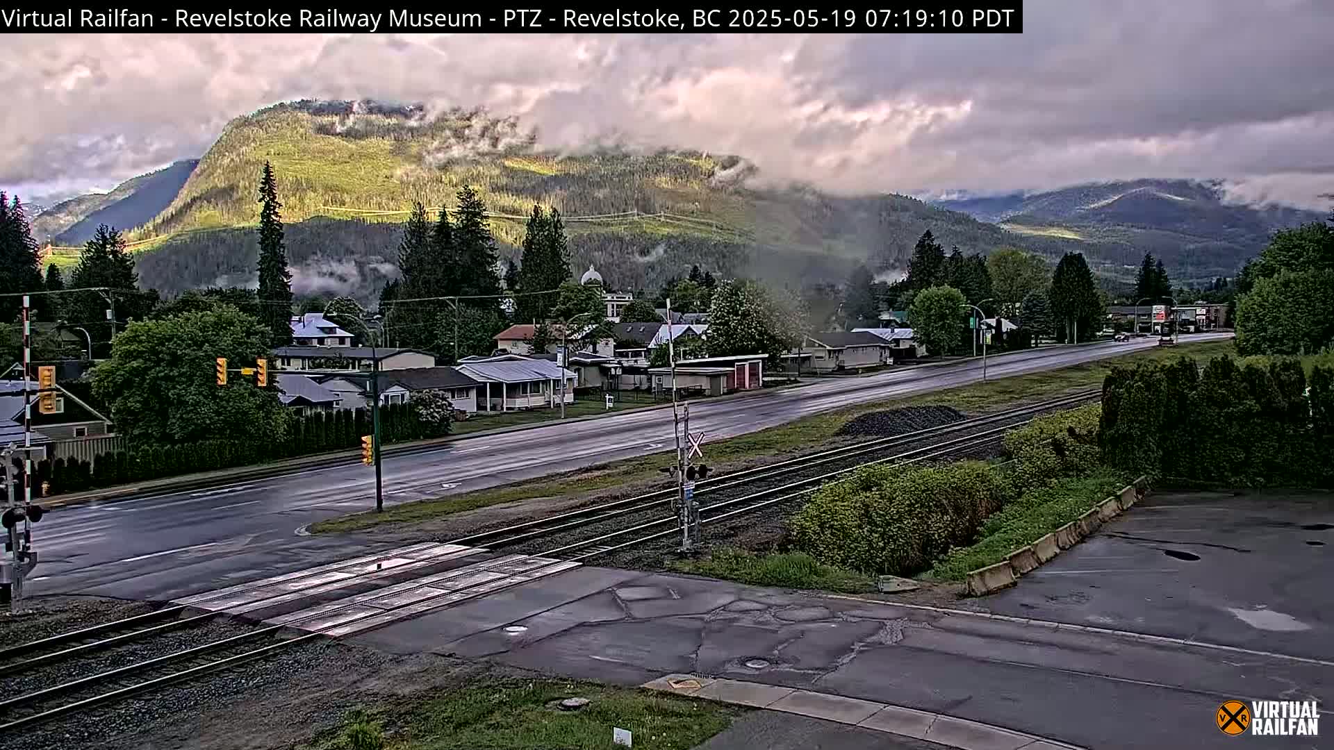 A wet road and railway tracks intersect in front of a small town nestled at the base of a mountain range under an overcast sky.