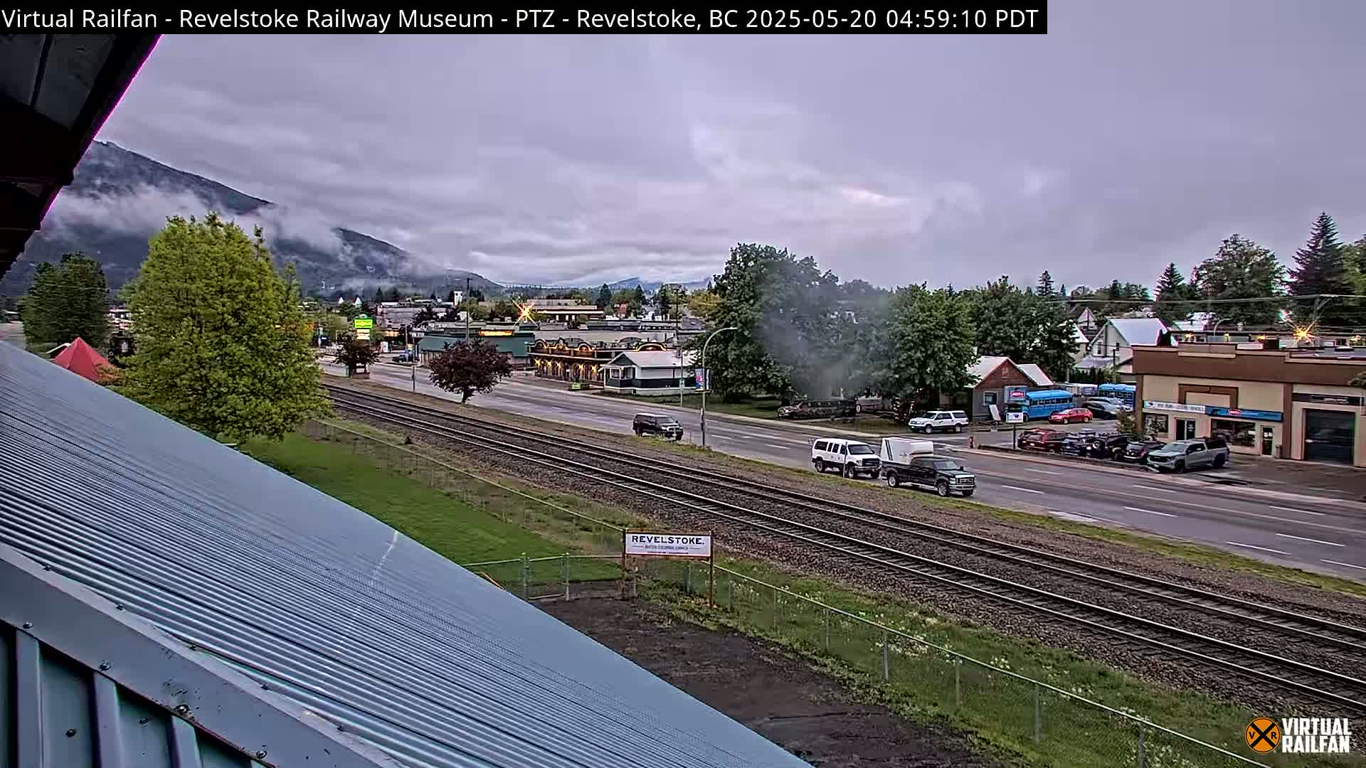 A cloudy, overcast view of a town with mountains in the background, featuring railway tracks running alongside a road with several vehicles, and a partially visible corrugated metal roof in the foreground.