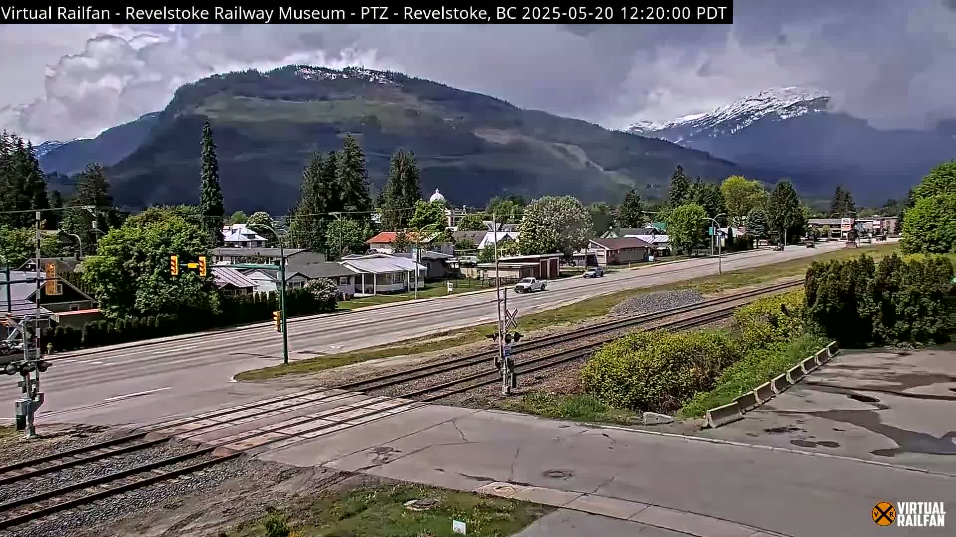 A partly cloudy day reveals a small town nestled in a valley, with mountains in the background, some of which are snow-capped; train tracks run through the town.