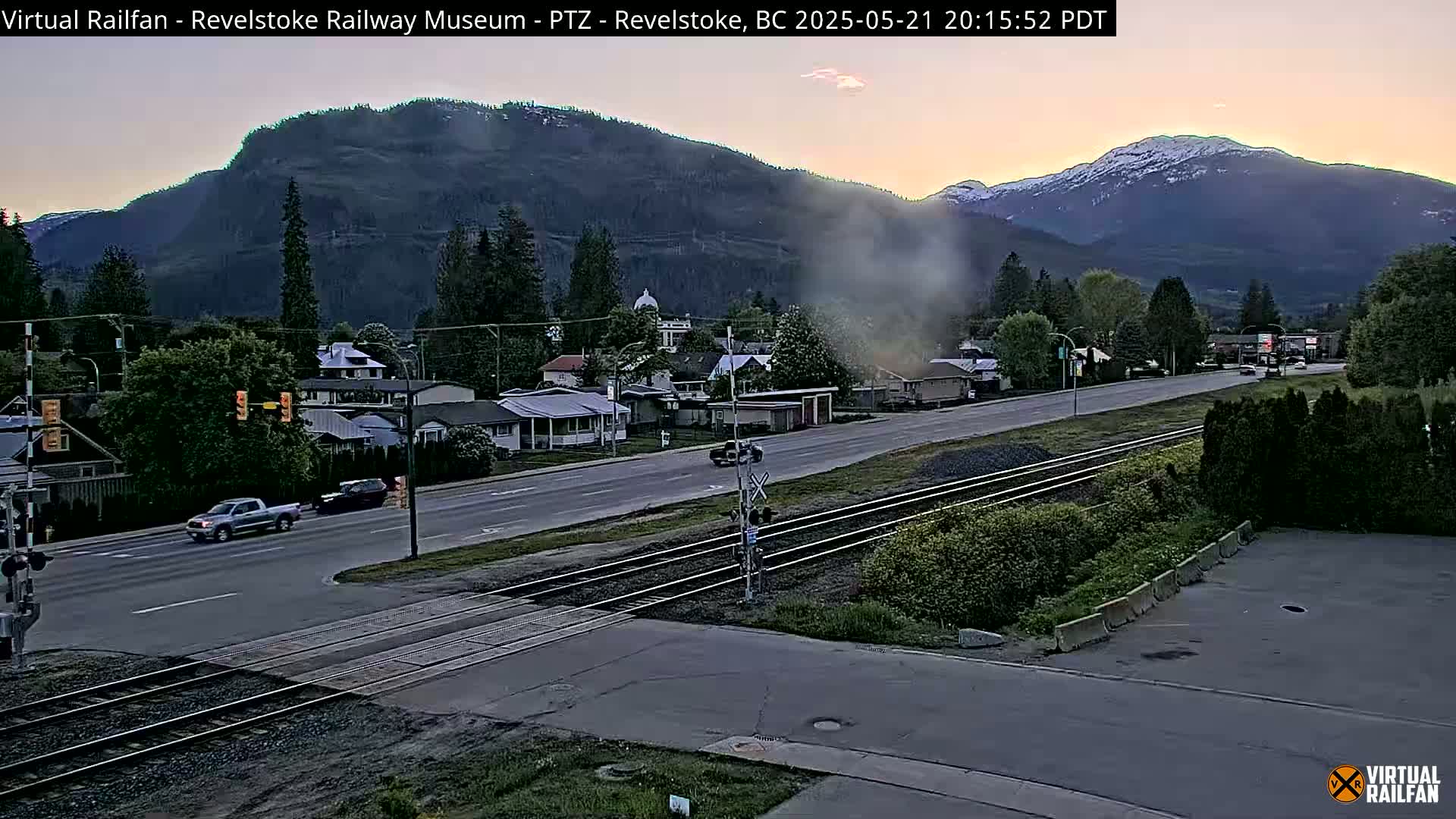 A twilight view of a town nestled in a valley, with a mountain range in the background showing some snow on the peaks, features a road crossing train tracks with several cars visible.