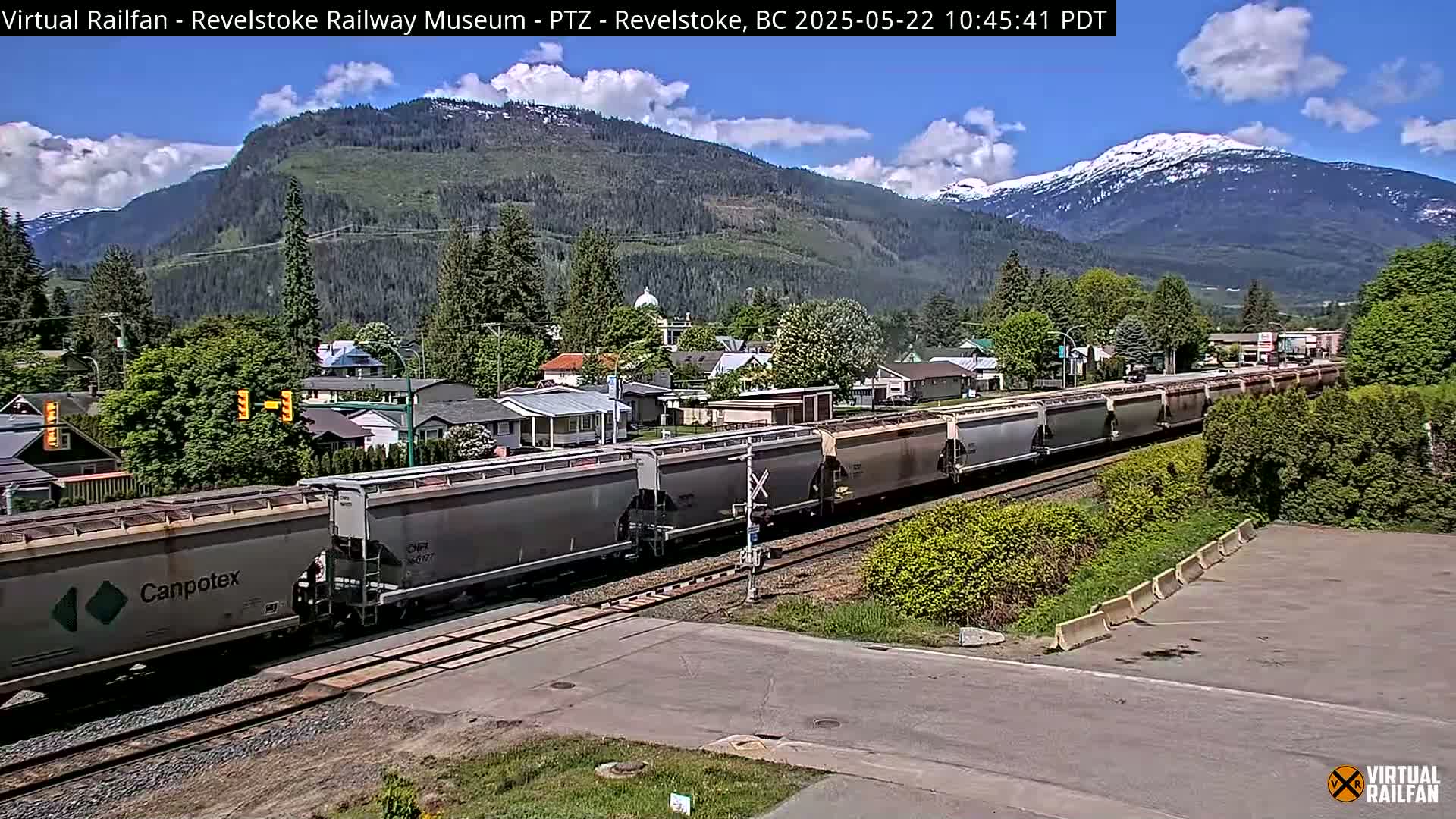A long freight train passes through a small town nestled in a valley, with mountains in the background under a partly cloudy sky.