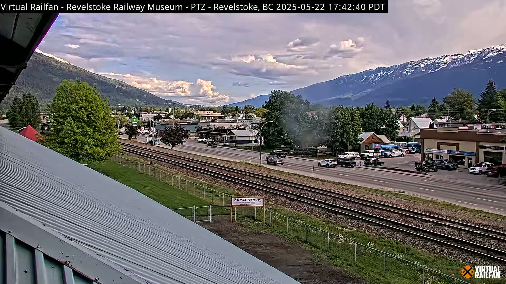A partly cloudy afternoon scene shows a small town nestled in a valley between mountains, with train tracks running through the center and cars parked along the road.